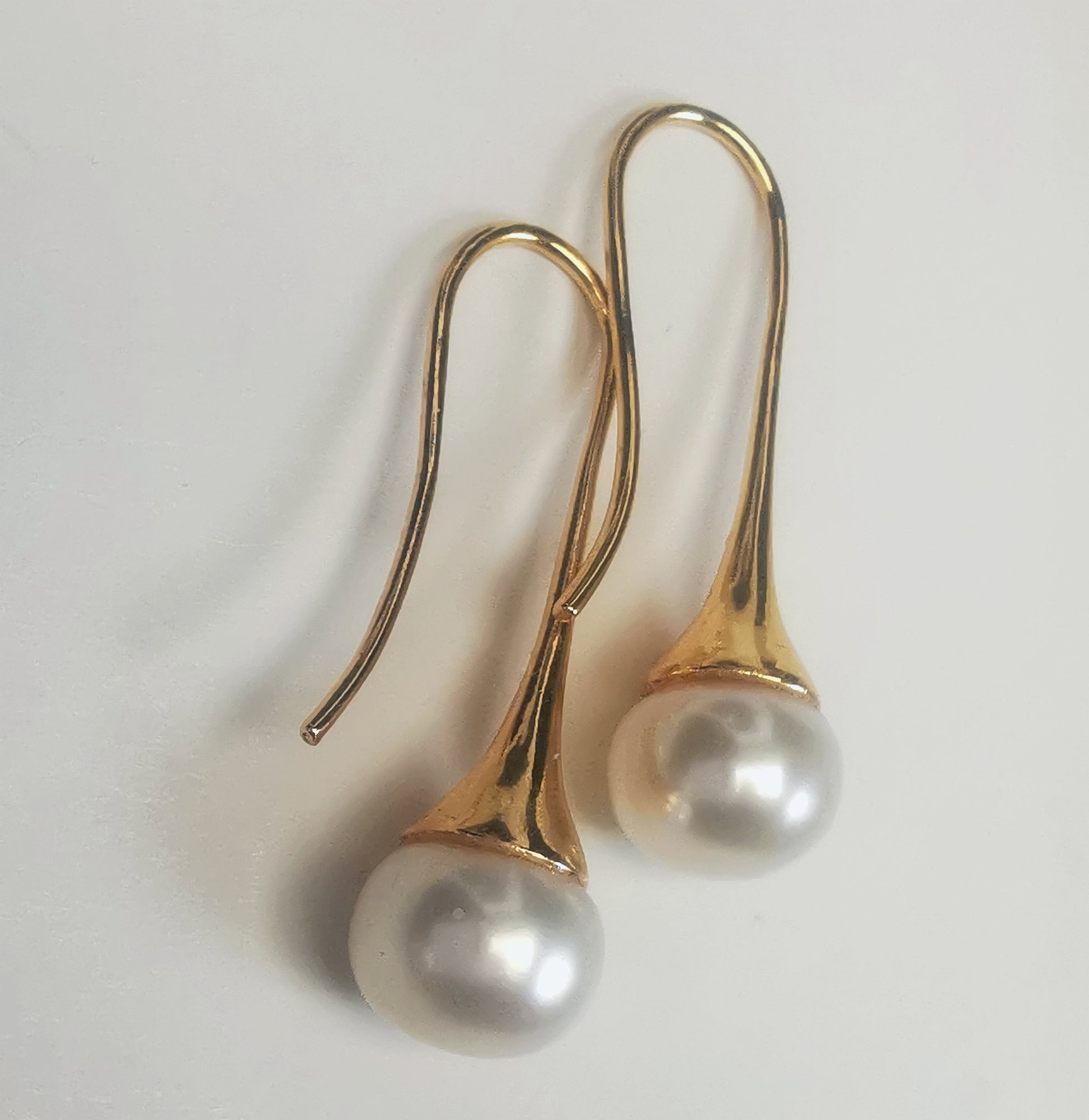 Boucles d'oreilles perles de culture blanches 9mm sur pendants longs plaqués or. Présentées cote o cote, perles en bas, pendants tournés vers la gauche, boucle droite décalée vers le haut, sur fond blanc. Vue en gros plan de dessus.