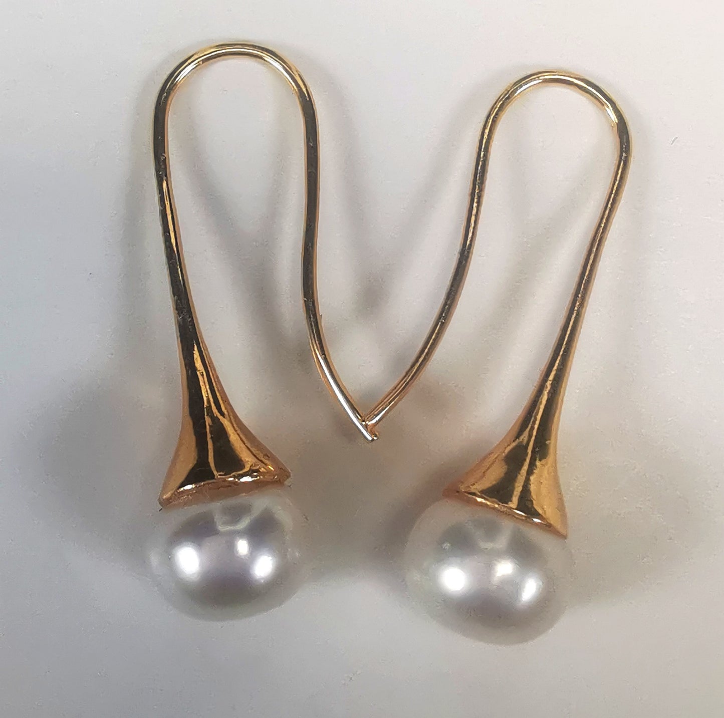 Boucles d'oreilles perles de culture blanches 9mm sur pendants longs plaqués or. Présentées cote o cote, perles en bas, boucle de gauche, pendant tourné vers la droite, celle de droite vers la gauche, sur fond blanc. Vue de dessus.