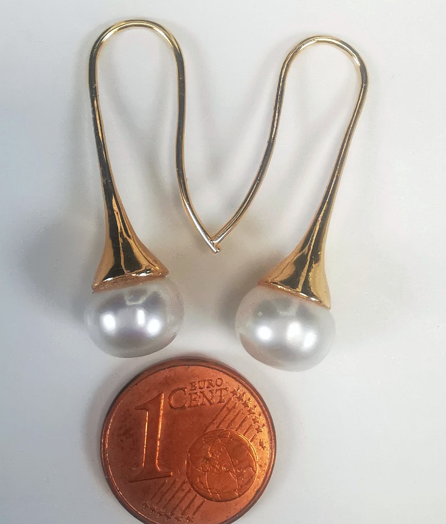 Boucles d'oreilles perles de culture blanches 9mm sur pendants longs plaqués or. Présentées cote o cote, perles en bas, boucle de gauche, pendant tourné vers la droite, celle de droite vers la gauche, au dessus d'une pièce de 1 centime, sur fond blanc. Vue de dessus.