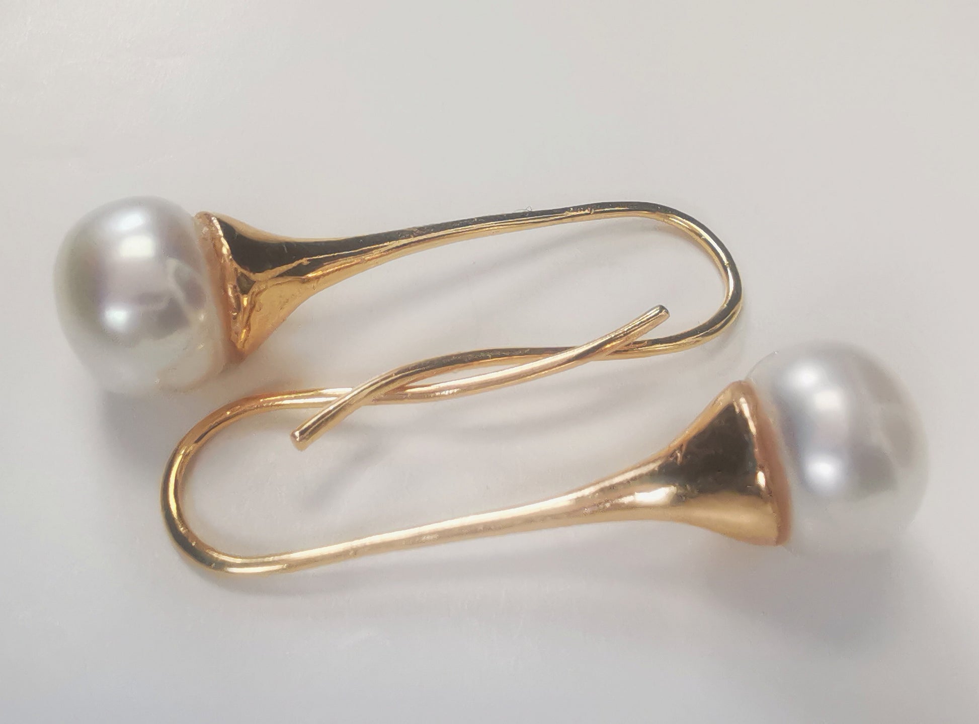 Boucles d'oreilles perles de culture blanches 9mm sur pendants longs plaqués or. Présentées horizontalement, boucle du dessus, perle à gauche pendant en bas, celle de dessous perle à droite pendant en haut, sur fond blanc. Vue de dessus.