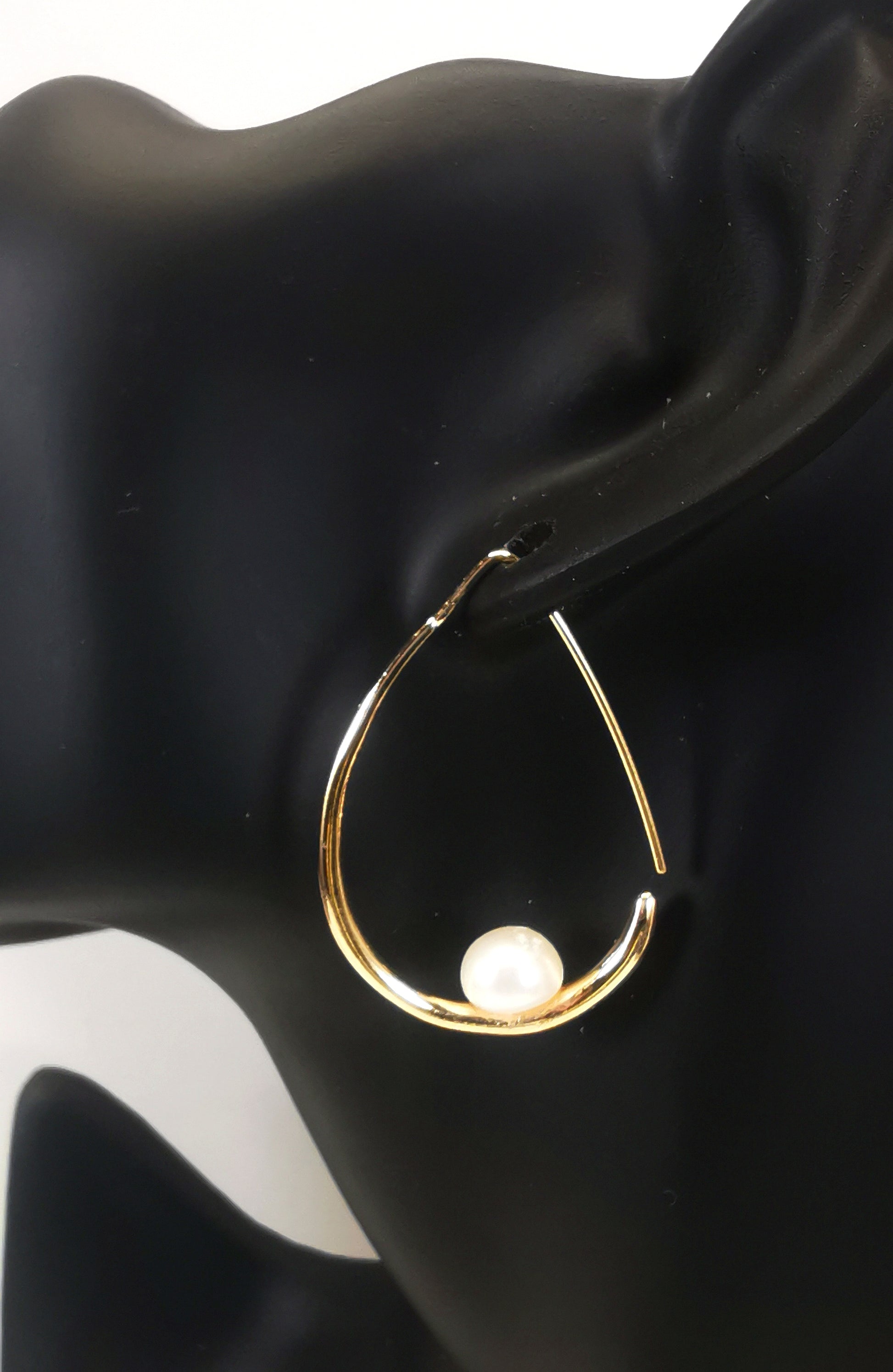 Boucles d'oreilles perles de culture blanches 8mm montées sur un pendant plaqué or. Ces pendant ont la forme d'un contour de poire de 40mm de haut, plus épais en bas avec un brin arrière fin pour pouvoir être enfilé sur l'oreille. Représentées par une boucle suspendue à l'oreille d'un mannequin buste noir. Vue de coté.