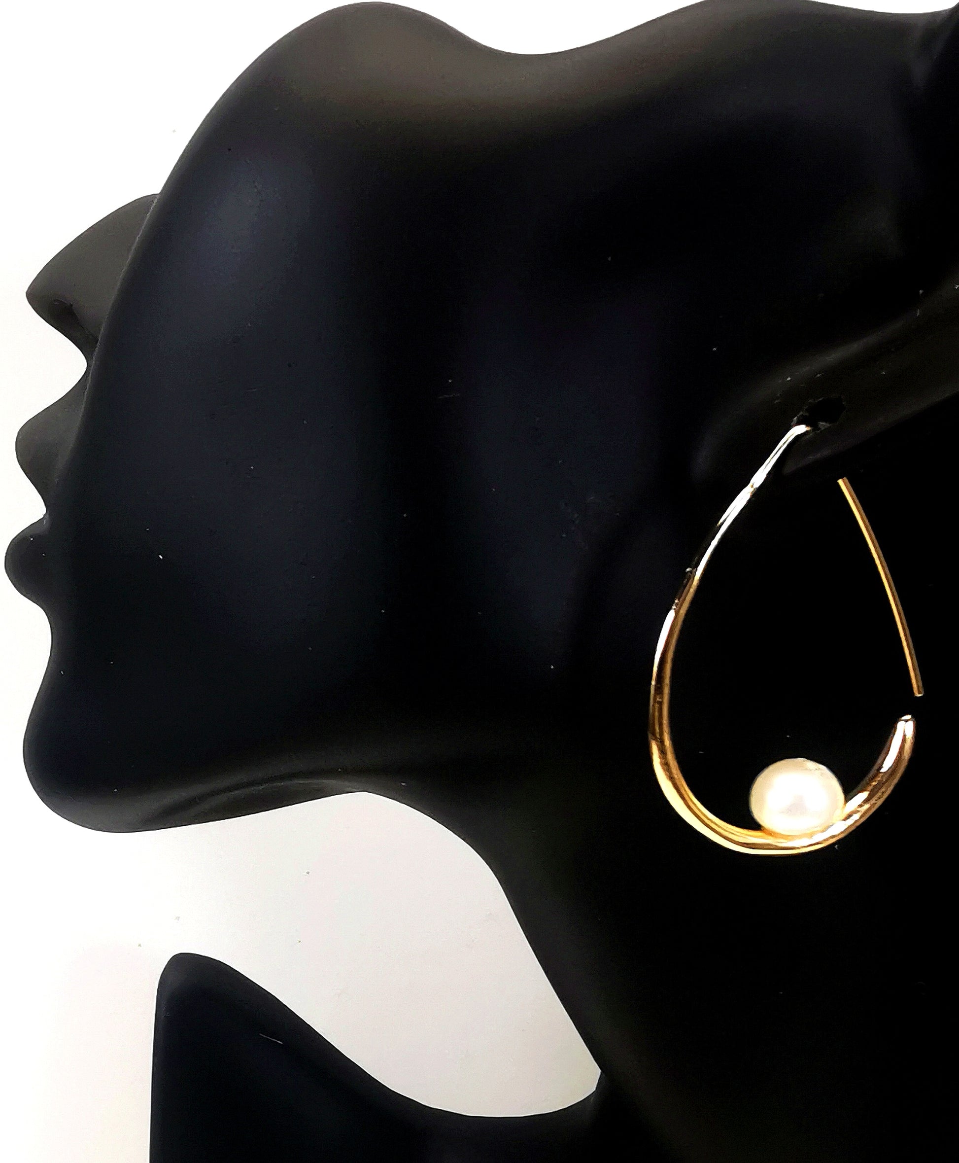 Boucles d'oreilles perles de culture blanches 8mm montées sur un pendant plaqué or. Ces pendants ont la forme d'un contour de poire de 40mm de haut, plus épais en bas avec un brin arrière fin pour pouvoir être enfilé sur l'oreille. Représentées par une boucle suspendue à l'oreille d'un mannequin buste noir. Vue en plan large de coté.