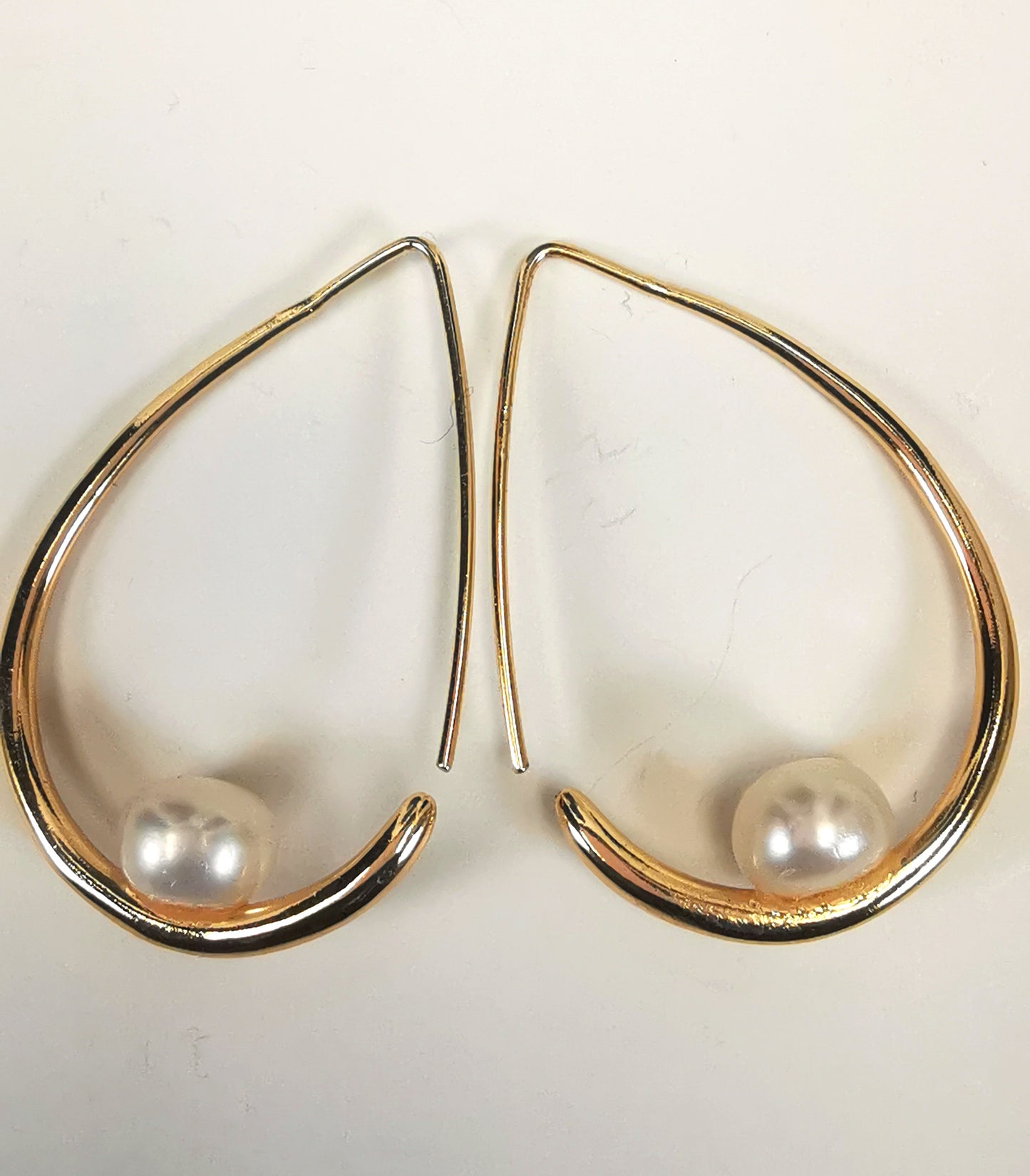 Boucles d'oreilles perles de culture blanches 8mm montées sur un pendant plaqué or. Ces pendants ont la forme d'un contour de poire de 40mm de haut, plus épais en bas avec un brin arrière fin pour pouvoir être enfilé sur l'oreille. Présentés posées cote à cote, en sens inverse, sur un fond blanc. Vue de dessus.