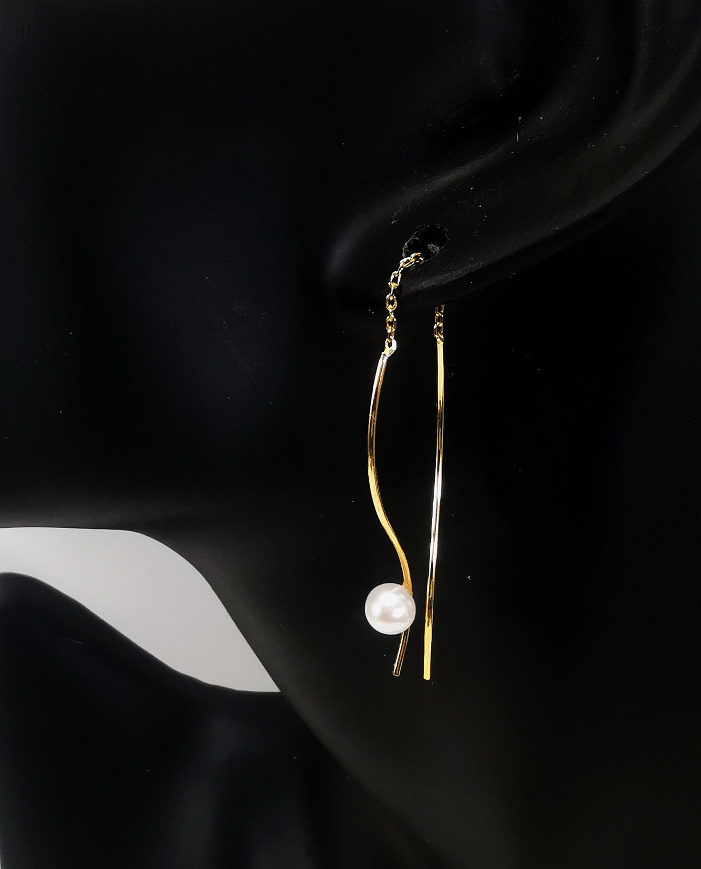 Boucles d'oreilles perles de culture blanches rondes 5mm sur pendants plaqués or. Les pendants sont constitués de deux "s" longs de 45mm reliés par 15mm de chaine. Sur un des 2 "s", la perle est montée perpendiculairement, à 8mm du bas. Représentées par une boucle montée sur l'oreille d'un mannequin buste noir, le "s" avec la perle devant le lobe, l'autre derrière, à la même hauteur. Vue de coté en très gros plan.