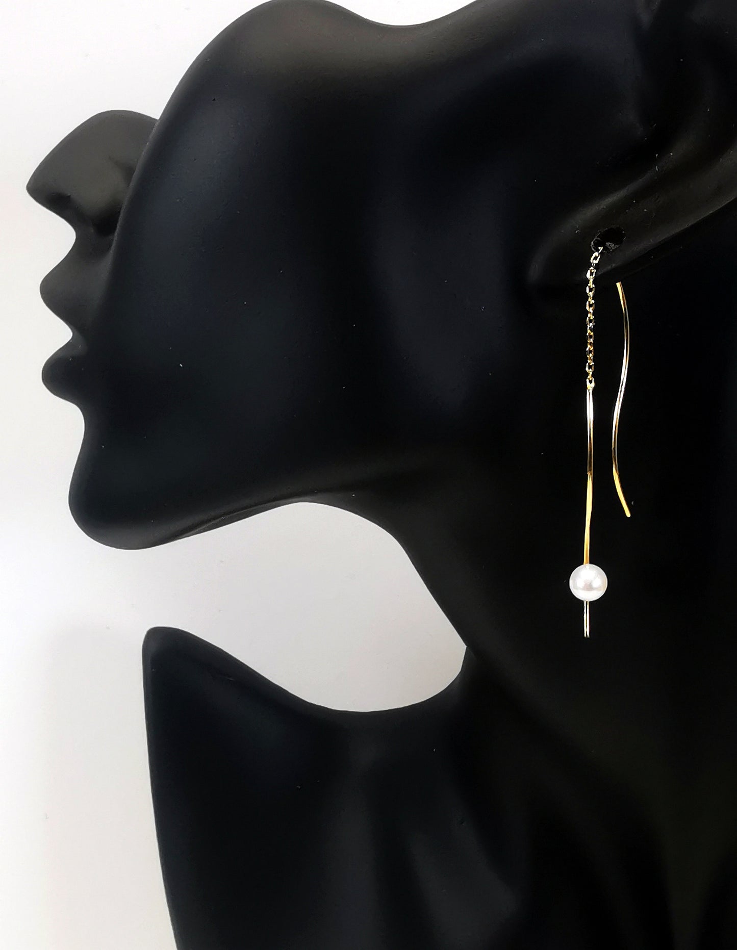 Boucles d'oreilles perles de culture blanches rondes 5mm sur pendants plaqués or. Les pendants sont constitués de deux "s" longs de 45mm reliés par 15mm de chaine. Sur un des 2 "s", la perle est montée perpendiculairement, à 8mm du bas. Représentées par une boucle montée sur l'oreille d'un mannequin buste noir, le "s" avec la perle devant le lobe et plus basse, l'autre derrière. Vue de coté.