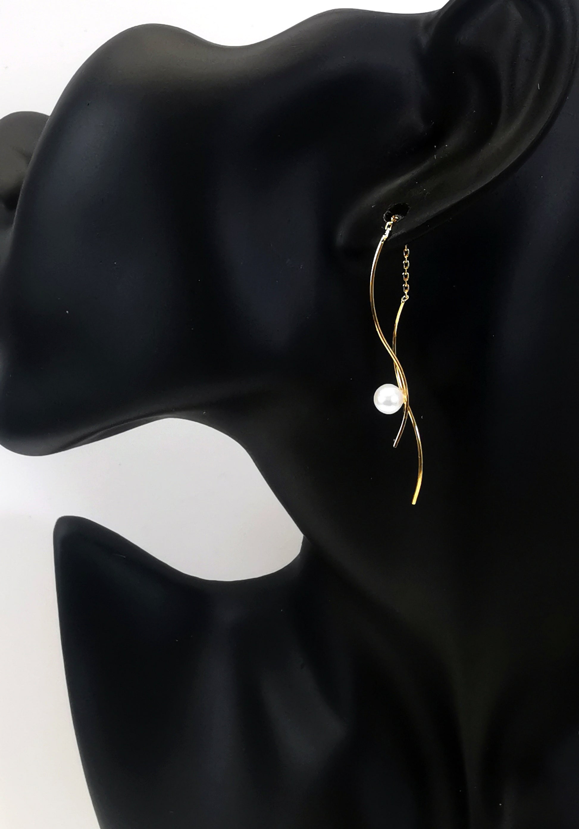 Boucles d'oreilles perles de culture blanches rondes 5mm sur pendants plaqués or. Les pendants sont constitués de deux "s" longs de 45mm reliés par 15mm de chaine. Sur un des 2 "s", la perle est montée perpendiculairement, à 8mm du bas. Représentées par une boucle montée sur l'oreille d'un mannequin buste noir,  le "s" avec la perle devant le lobe, l'autre derrière. Vue de coté en plan large.