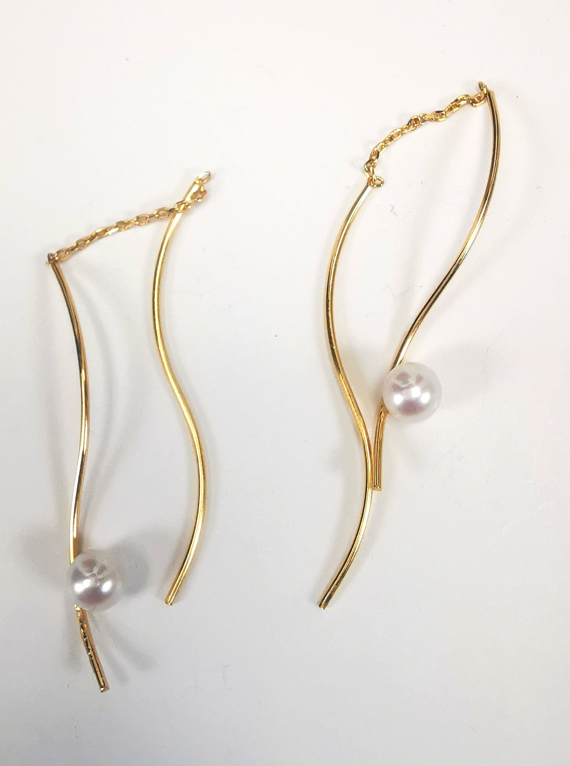 Boucles d'oreilles perles de culture blanches rondes 5mm sur pendants plaqués or. Les pendants sont constitués de deux "s" longs de 45mm reliés par 15mm de chaine. Sur un des 2 "s", la perle est montée perpendiculairement, à 8mm du bas. Présentées cote à cote, perle en bas, boucle de droite perle à droite, celle de gauche, perle à gauche, posées sur fond blanc. Vue de dessus.
