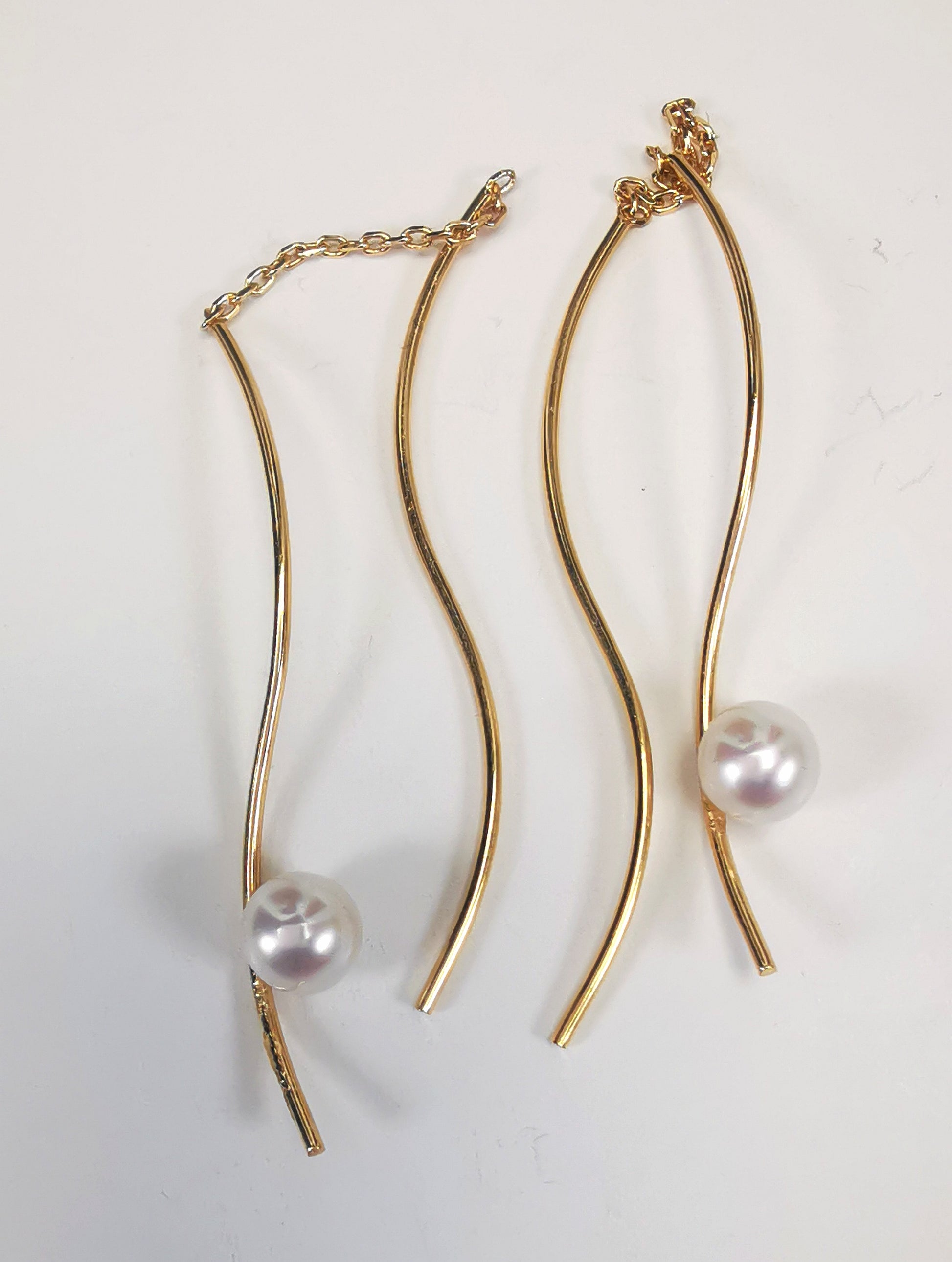 Boucles d'oreilles perles de culture blanches rondes 5mm sur pendants plaqués or. Les pendants sont constitués de deux "s" longs de 45mm reliés par 15mm de chaine. Sur un des 2 "s", la perle est montée perpendiculairement, à 8mm du bas. Présentées cote à cote, perle en bas, la boucle de droite décalée vers le haut,  posées sur fond blanc. Vue de dessus.