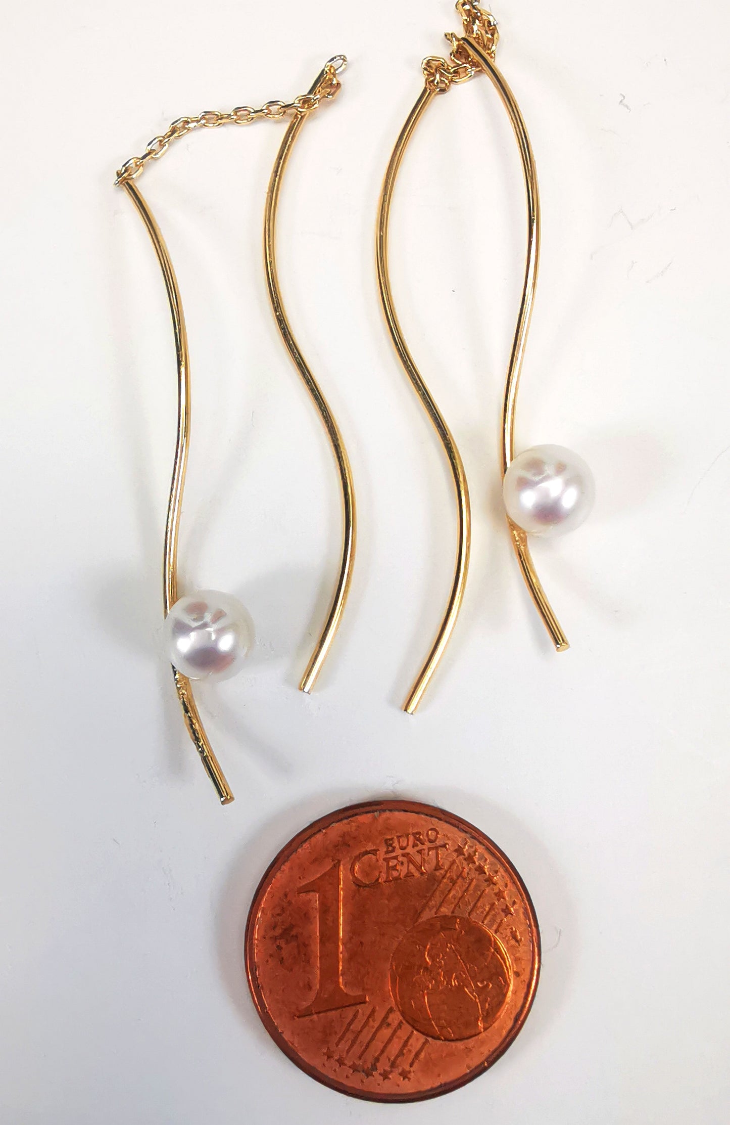 Boucles d'oreilles perles de culture blanches rondes 5mm sur pendants plaqués or. Les pendants sont constitués de deux "s" longs de 45mm reliés par 15mm de chaine. Sur un des 2 "s", la perle est montée perpendiculairement, à 8mm du bas. Présentées cote à cote, perle en bas, au dessus d'une pièce de 1 centime, posées sur fond blanc. Vue de dessus.