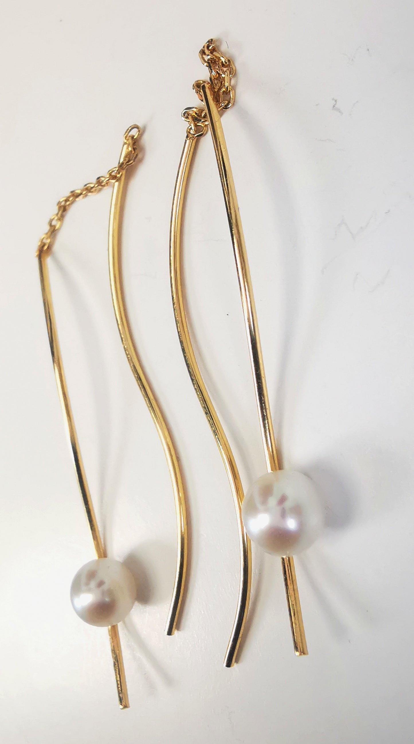 Boucles d'oreilles perles de culture blanches rondes 5mm sur pendants plaqués or. Les pendants sont constitués de deux "s" longs de 45mm reliés par 15mm de chaine. Sur un des 2 "s", la perle est montée perpendiculairement, à 8mm du bas. Présentées cote à cote, perle en bas, posées sur fond blanc. Vue en gros plan depuis la droite.
