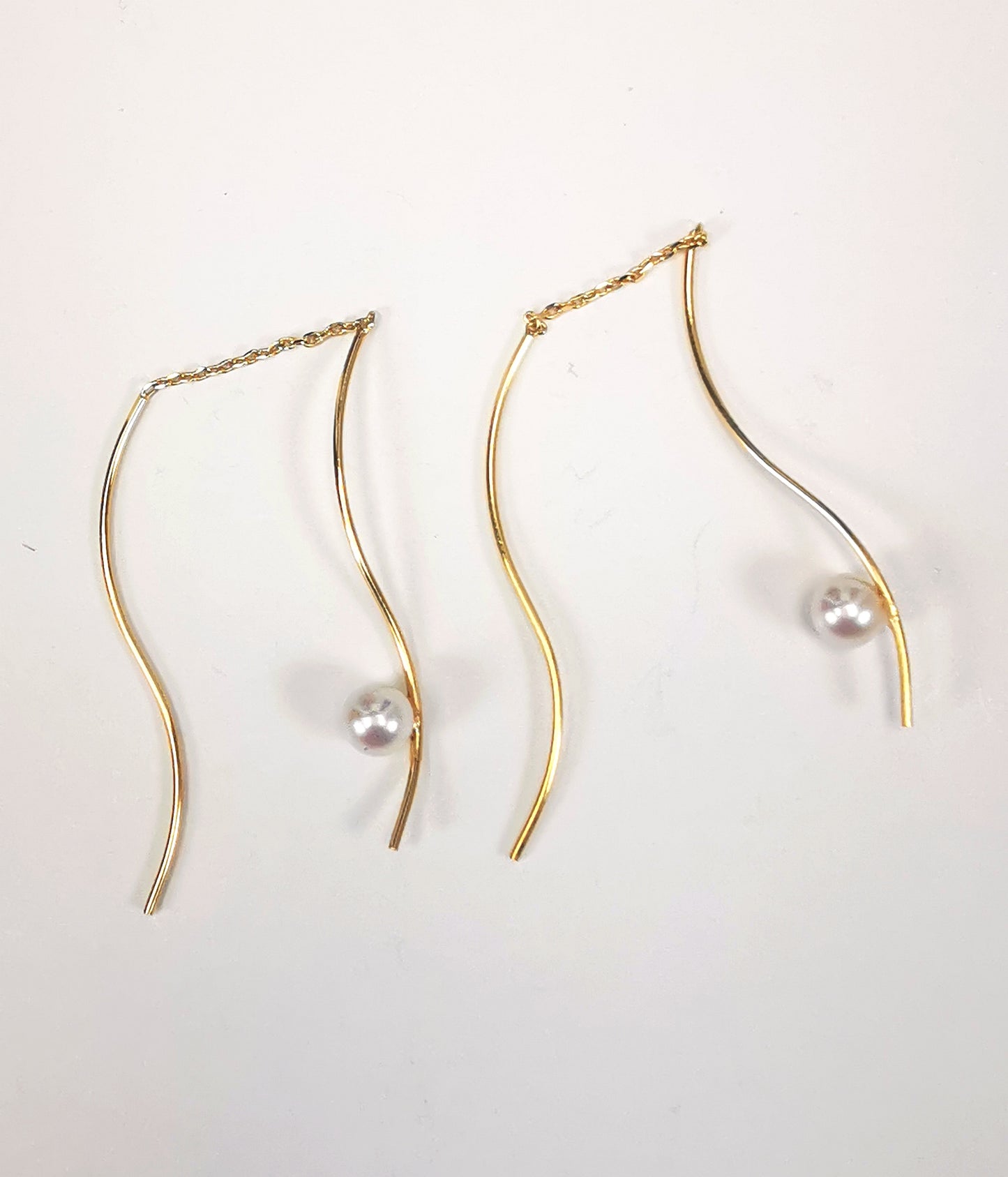 Boucles d'oreilles perles de culture blanches rondes 5mm sur pendants plaqués or. Les pendants sont constitués de deux "s" longs de 45mm reliés par 15mm de chaine. Sur un des 2 "s", la perle est montée perpendiculairement, à 8mm du bas. Présentées cote à cote, perle en bas,  tournées vers la gauche, posées sur fond blanc. Vue de dessus.