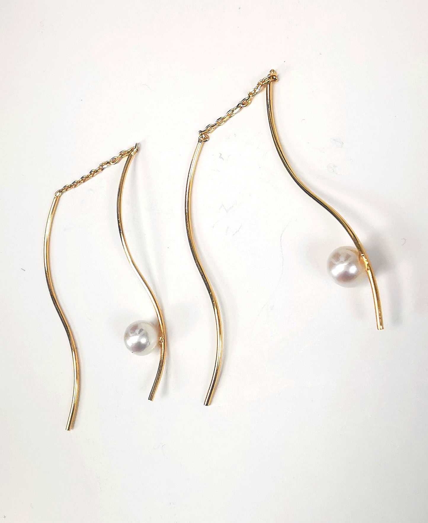 Boucles d'oreilles perles de culture blanches rondes 5mm sur pendants plaqués or. Les pendants sont constitués de deux "s" longs de 45mm reliés par 15mm de chaine. Sur un des 2 "s", la perle est montée perpendiculairement, à 8mm du bas. Présentées cote à cote, perle en bas, tournées vers la gauche, la boucle de droite décalée vers le haut, posées sur fond blanc. Vue de dessus.
