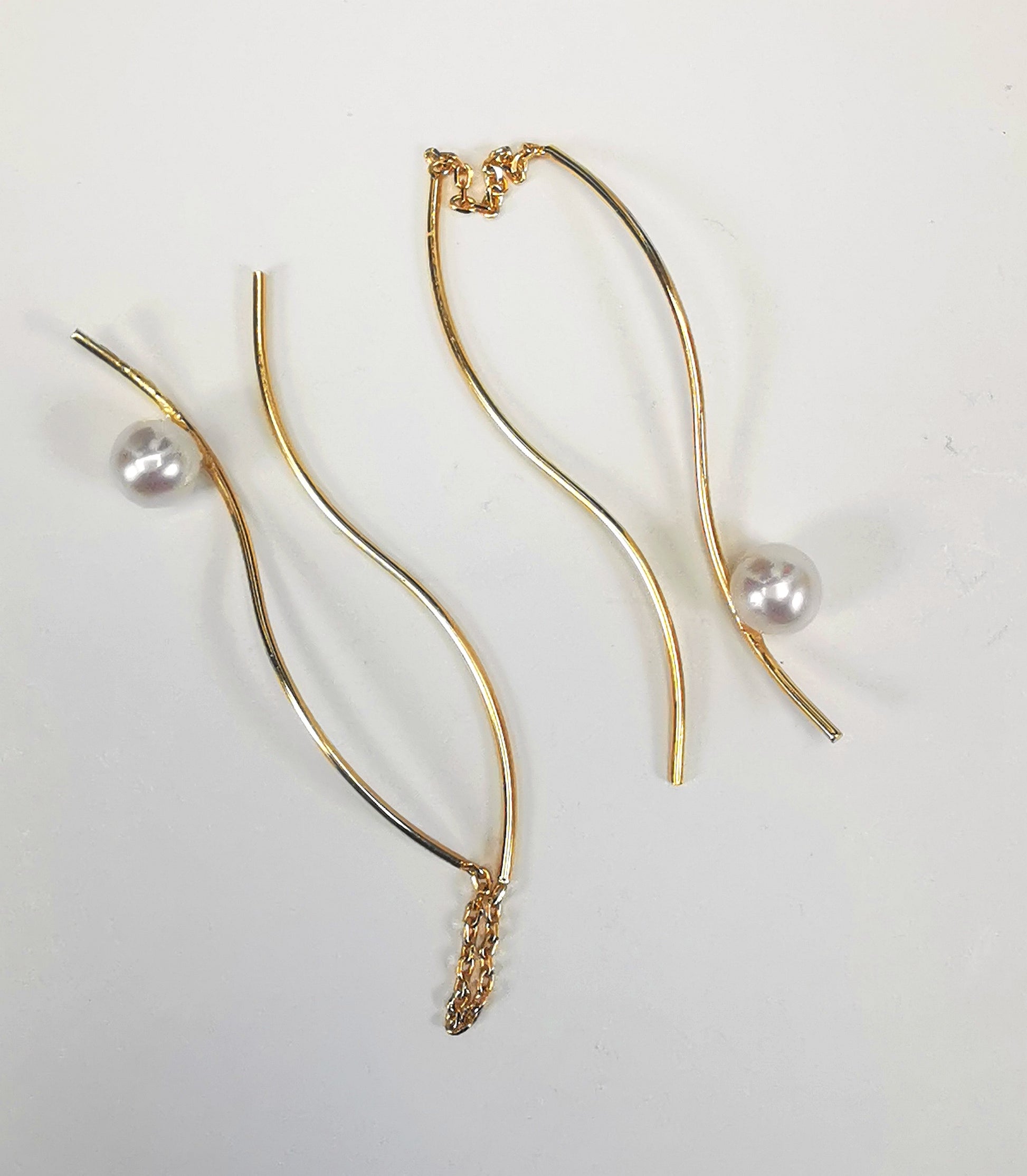 Boucles d'oreilles perles de culture blanches rondes 5mm sur pendants plaqués or. Les pendants sont constitués de deux "s" longs de 45mm reliés par 15mm de chaine. Sur un des 2 "s", la perle est montée perpendiculairement, à 8mm du bas. Présentées cote à cote, boucle de gauche perle en haut, celle de droite perle en bas, , posées sur fond blanc. Vue de dessus.