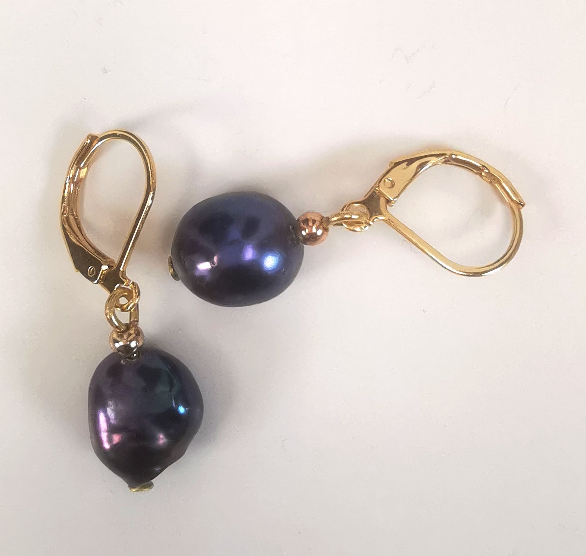 Boucles d'oreilles perles de culture noires baroques 10-12mm montées sur dormeuses plaquées or. Présentées perpendiculairement, celle de gauche verticale perles en bas, celle de droite horizontale, perle à gauche, sur un fond blanc. Vue de dessus.