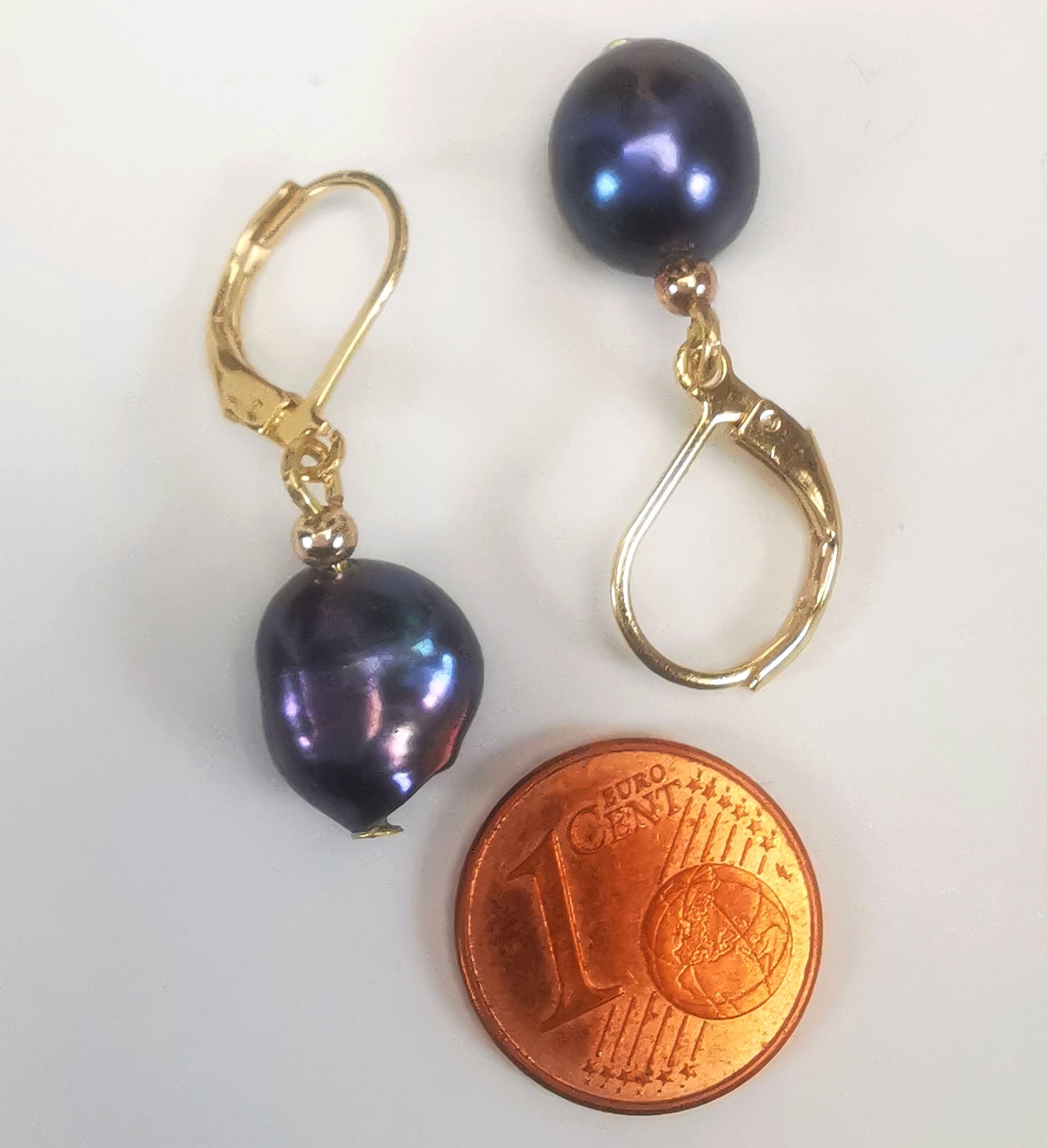 Boucles d'oreilles perles de culture noires baroques 10-12mm montées sur dormeuses plaquées or. Présentées cote à cote, têtes bèches, boucle de droite perles en bas, celle de gauche perle en haut, au dessus d'une pièce de 1 centime, sur un fond blanc. Vue de dessus.