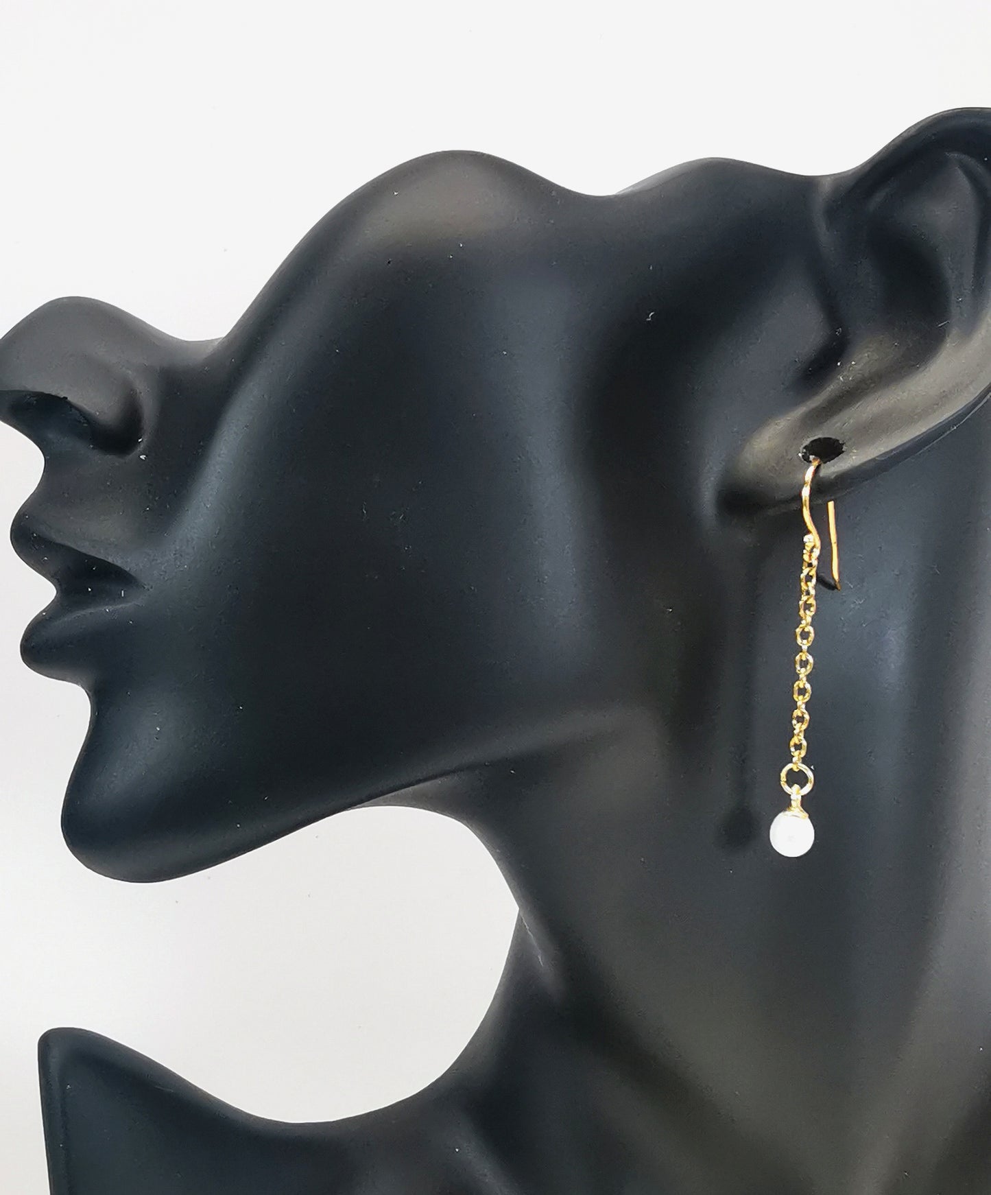 Boucles d'oreilles perles de culture blanches rondes 5mm sur pendants et chaînes plaqués or. Représentées par une boucle accrochée à une oreille de mannequin buste noir. Vue de coté en plan large.