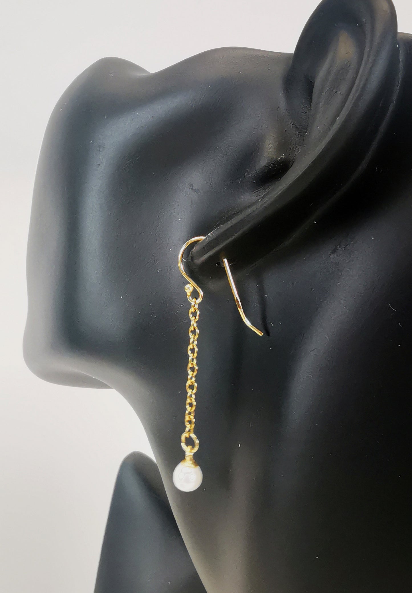 Boucles d'oreilles perles de culture blanches rondes 5mm sur pendants et chaînes plaqués or. Représentées par une boucle accrochée à une oreille de mannequin buste noir. Vue de trois quarts arrière.