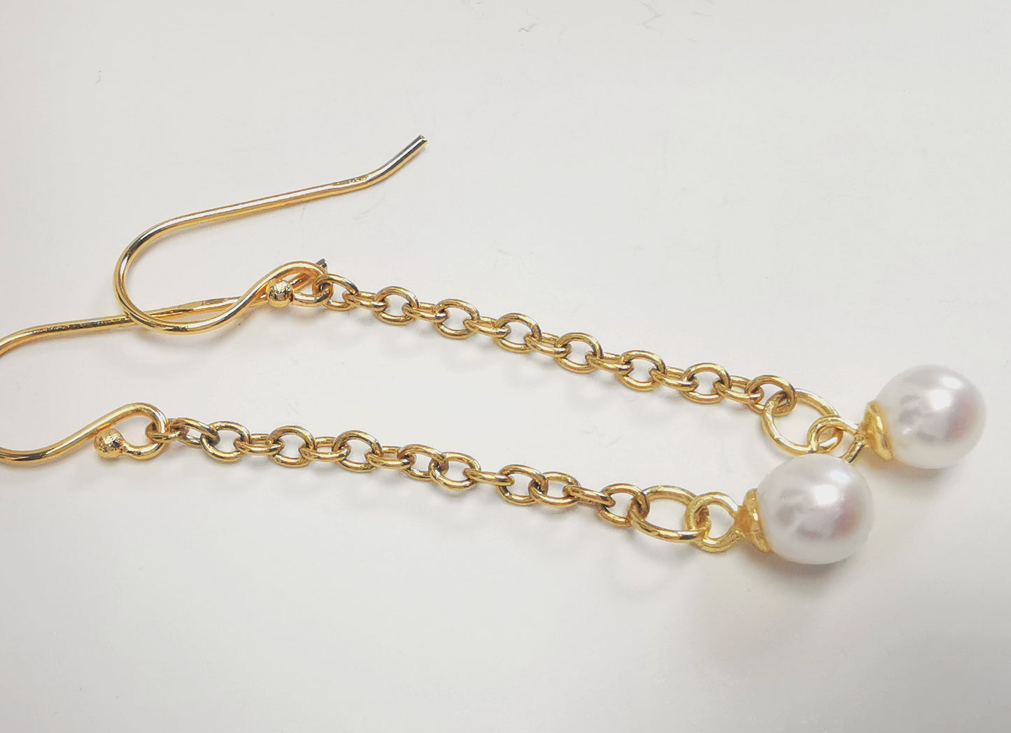 Boucles d'oreilles perles de culture blanches rondes 5mm sur pendants et chaînes plaqués or. Présentées à l'horizontale, l'une au dessus de l'autre, perles à droite, sur fond blanc. Vue de dessus.