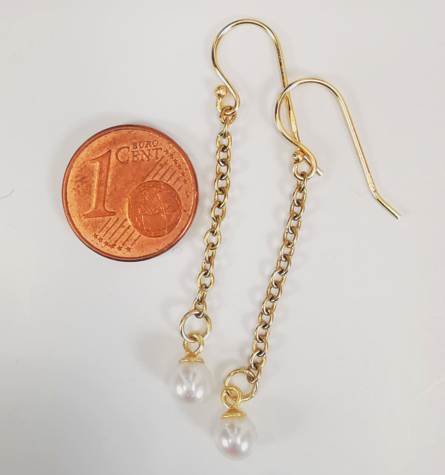 Boucles d'oreilles perles de culture blanches rondes 5mm sur pendants et chaînes plaqués or. Présentées cote à cote, boucle de gauche décalée vers le haut, à droite d'une pièce de 1 centime, sur fond blanc. Vue de dessus.