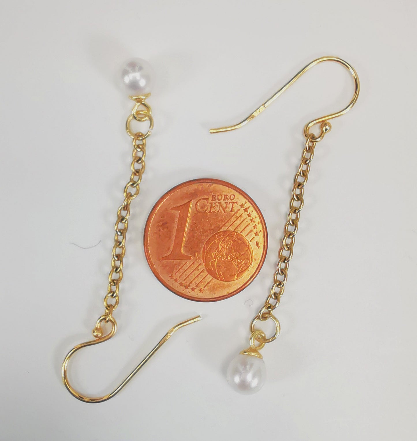 Boucles d'oreilles perles de culture blanches rondes 5mm sur pendants et chaînes plaqués or. Présentées cote à cote, têtes bèches, boucle de droite perle en haut, celle de droite perle en bas, sur fond blanc. Vue de dessus.