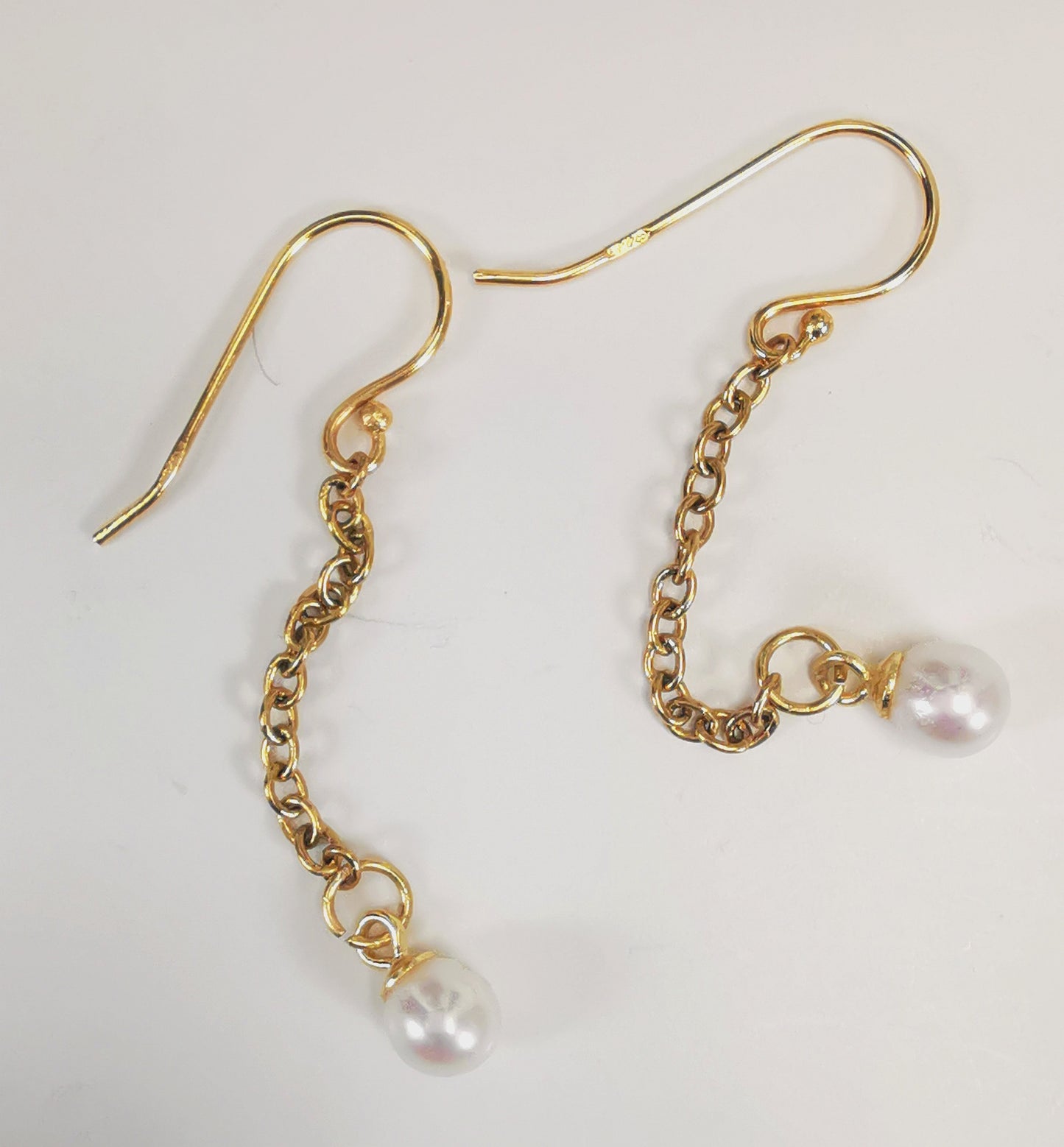 Boucles d'oreilles perles de culture blanches rondes 5mm sur pendants et chaînes plaqués or. Présentées cote à cote, boucle de gauche décalée vers le haut, les deux chaines détendues, sur fond blanc. Vue de dessus.