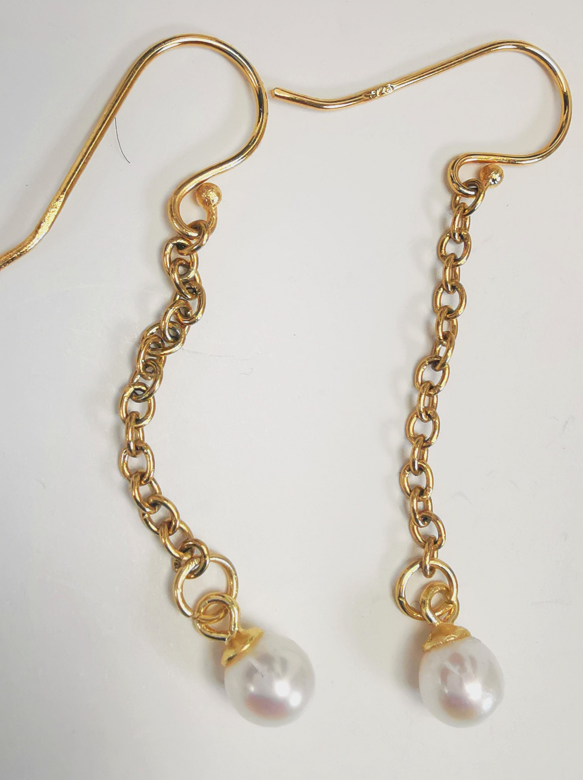Boucles d'oreilles perles de culture blanches rondes 5mm sur pendants et chaînes plaqués or. Présentées cote à cote, boucle de gauche chaîne détendue, sur fond blanc. Vue de dessus.