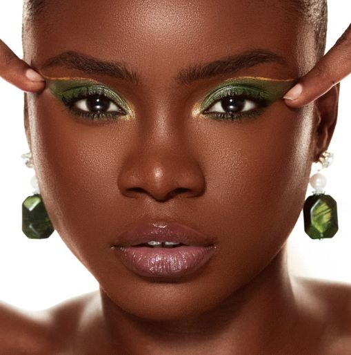 Visage en gros plan d'une très jolie jeune femme noire, aux yeux maquillés de vert, avec en évidence, ces boucles d'oreilles avec une perle pendante et en dessous une très grosse pierre verte rectangulaire.