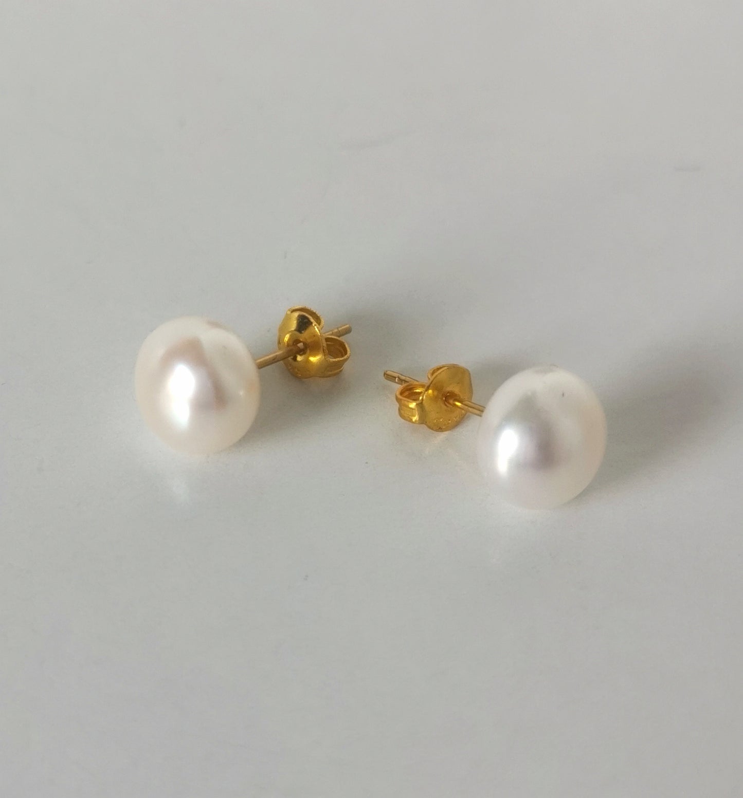Boucles d'oreilles perles de culture blanches 9mm montées sur clous plaqués or. Présentées cote à cote, perles en bas, boucle de gauche inclinée vers la gauche, celle de droite vers la droite, posées sur un fond blanc. Vue de face.