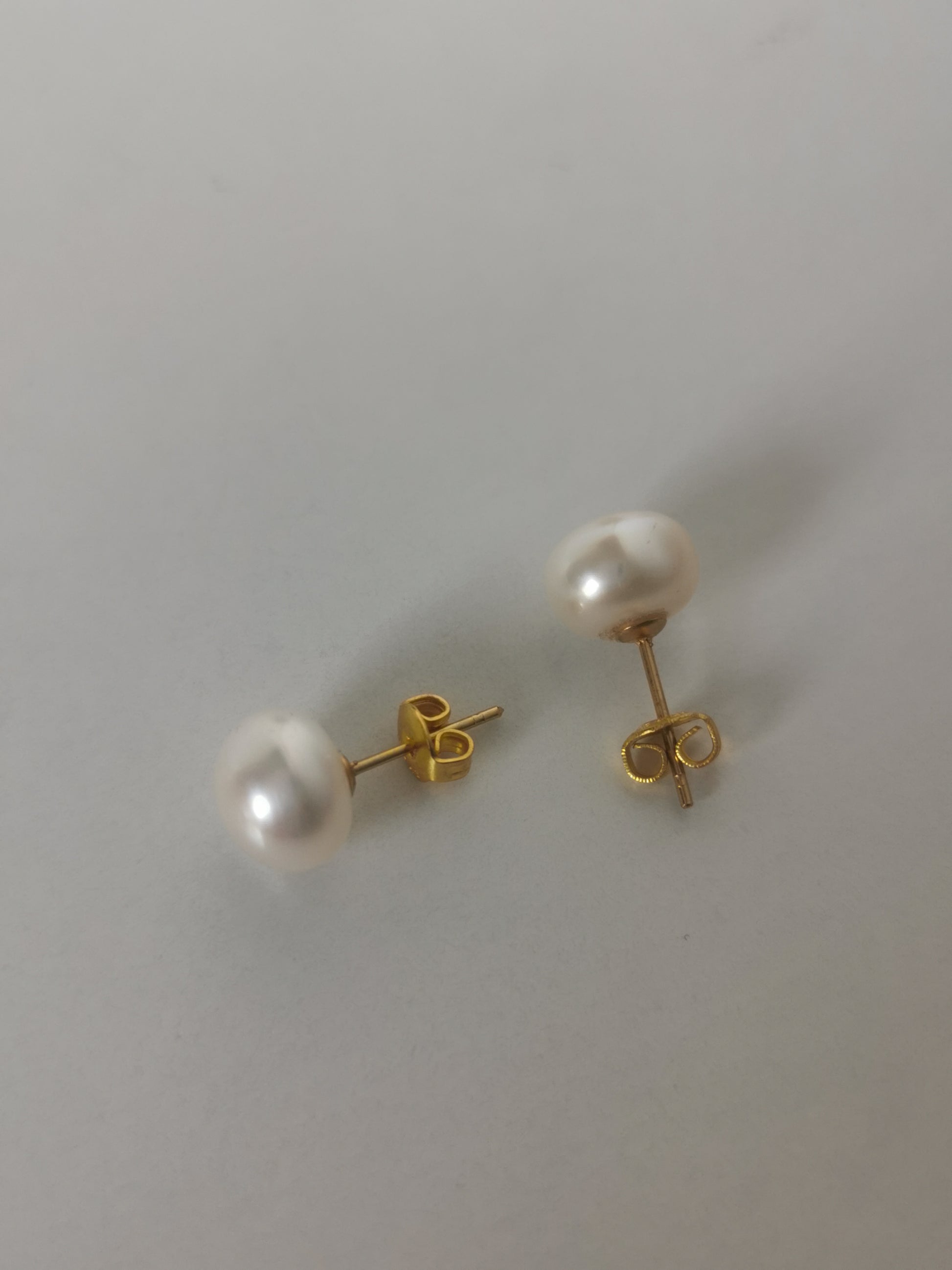 Boucles d'oreilles perles de culture blanches 9mm montées sur clous plaqués or. Présentées perpendiculairement, boucle de gauche à l'horizontale, perle à gauche, celle de droite verticalement perle en haut, posées sur un fond blanc. Vue de dessus.