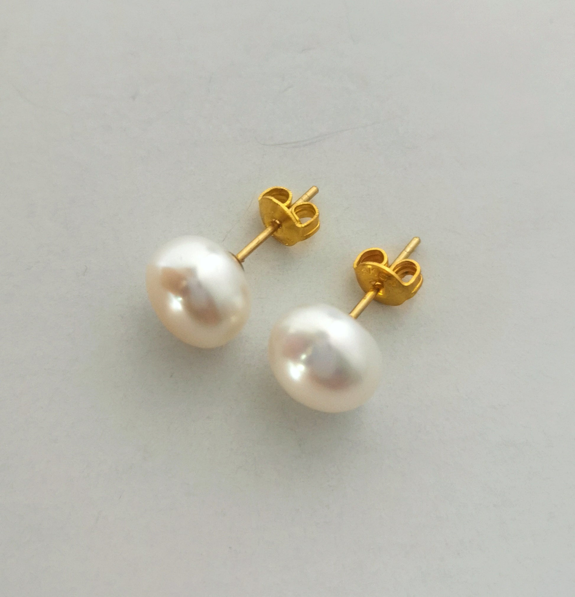 Boucles d'oreilles perles de culture blanches 9mm montées sur clous plaqués or. Présentées cote à cote, perles en bas, fermoirs inclinées vers la gauche, posées sur un fond blanc. Vue de face.