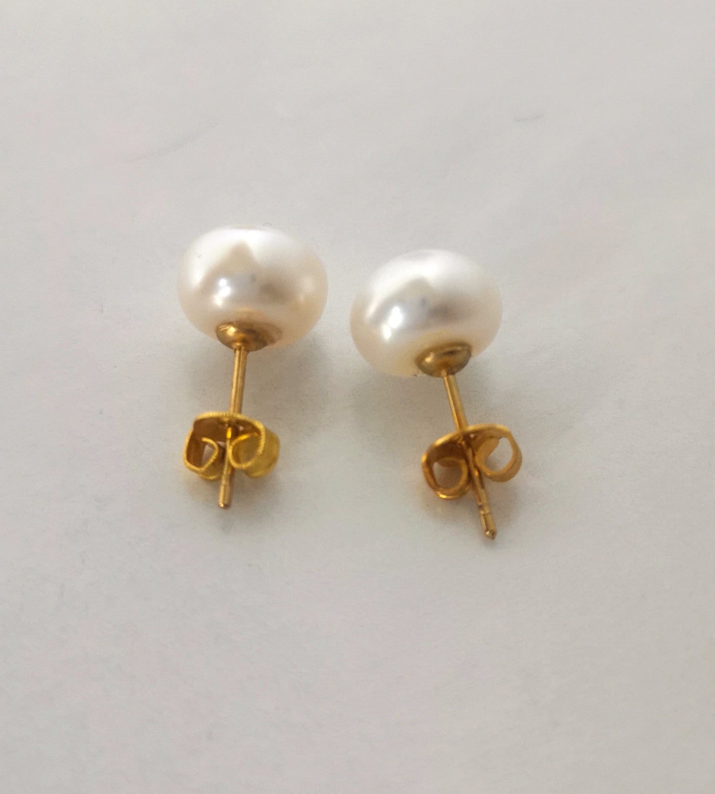 Boucles d'oreilles perles de culture blanches 9mm montées sur clous plaqués or. Présentées cote à cote, perles en haut, posées sur un fond blanc. Vue en très gros plan de dessus.