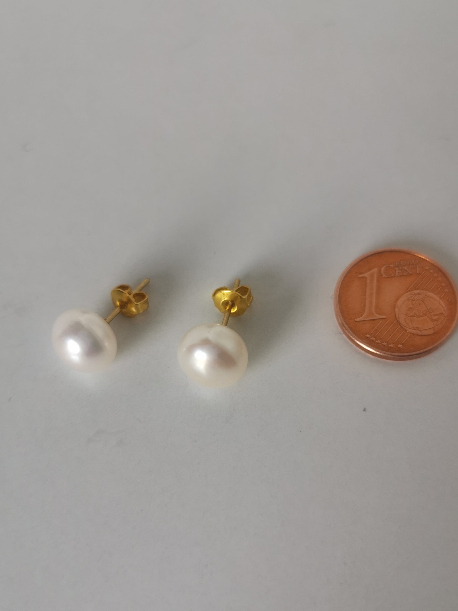 Boucles d'oreilles perles de culture blanches 9mm montées sur clous plaqués or. Présentées cote à cote, perles en bas, posées sur un fond blanc. A leur droite est posée une pièce de 1 centime. Vue de dessus.