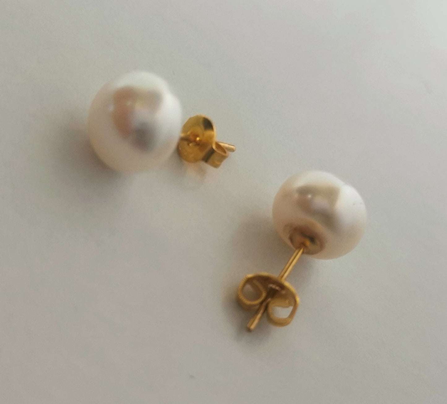 Boucles d'oreilles perles de culture blanches 9mm montées sur clous plaqués or. Présentées perpendiculairement, boucle de gauche perle à gauche, boucle de droite verticale, perle en haut, posées sur un fond blanc. Vue de dessus en gros plan.