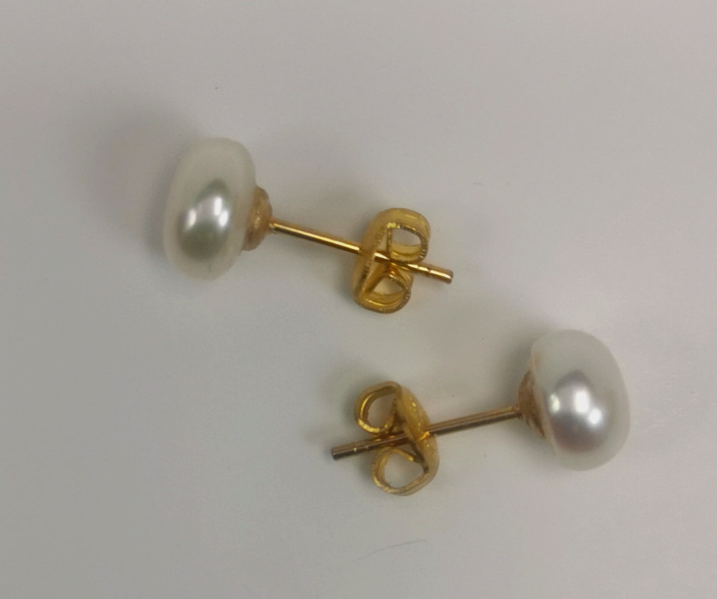 Boucles d'oreilles perles de culture blanches 9mm montées sur clous plaqués or. Présentées à l'horizontale, l'une au dessus de l'autre, boucle de dessus perle à gauche, celle de dessous perle à droite, posées sur un fond blanc. Vue de face.