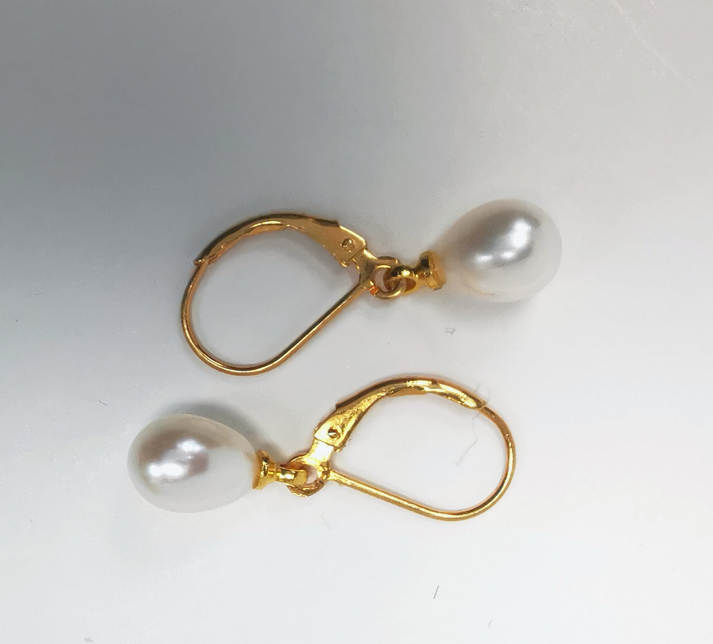Boucles d'oreilles perles de culture blanches 7-9mm sur dormeuses plaquées or. Présentées posées sur un fond blanc, à l'horizontale, celle de dessus perle à droite , celle de dessous perle à gauche. Vue de dessus.