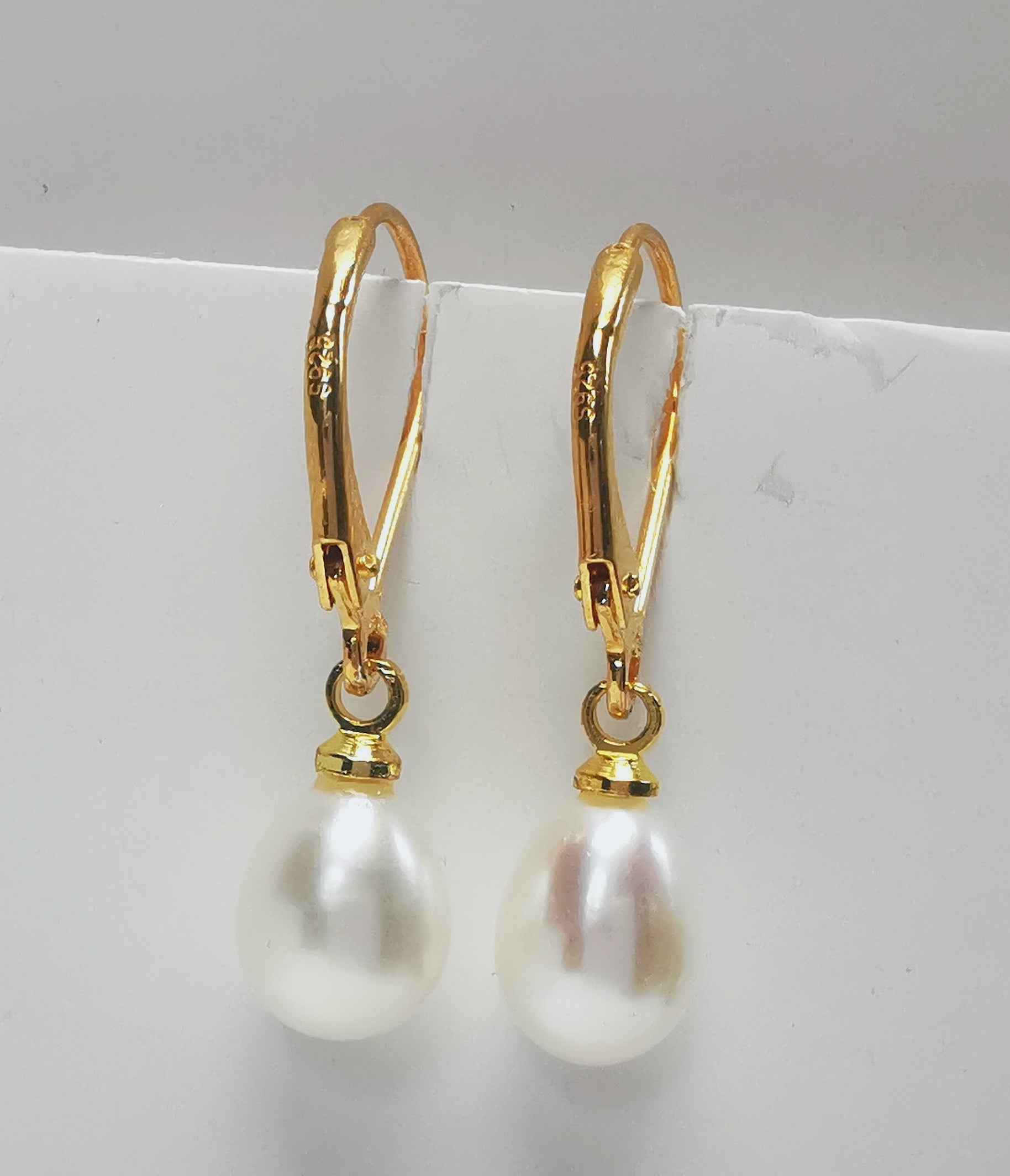 Boucles d'oreilles perles de culture blanches 7-9mm sur dormeuses plaquées or. Présentées suspendues à un support, cote à cote. Vue de face en gros plan.