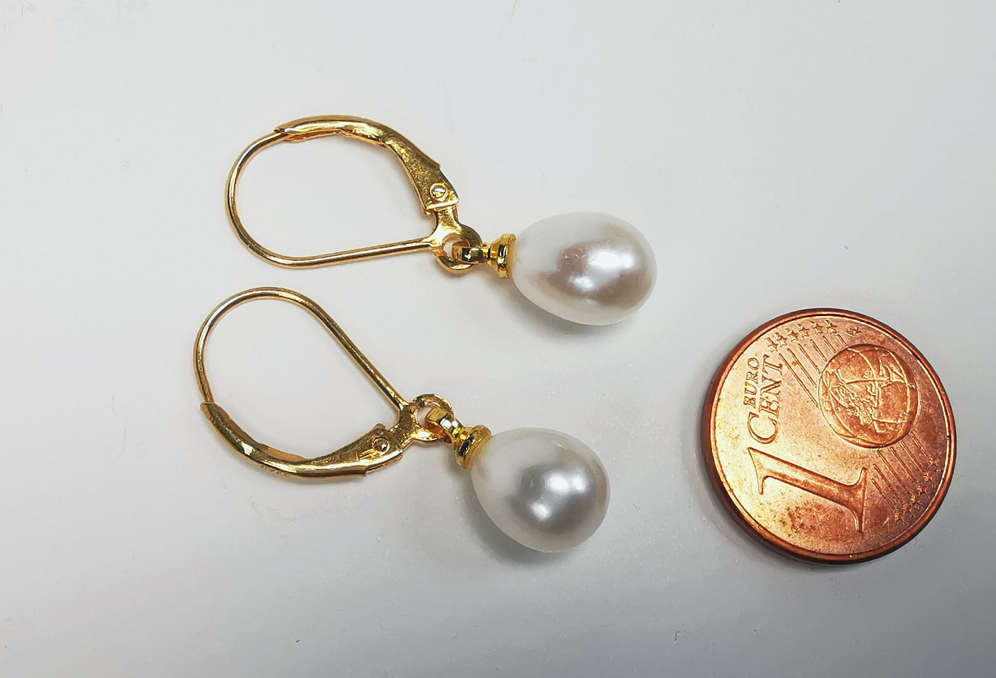 Boucles d'oreilles perles de culture blanches 7-9mm sur dormeuses plaquées or. Présentées posées sur un fond blanc, à l'horizontale, cote à cote, perles à gauche. Une pièce de un centime est posée à droite. Vue de dessus.