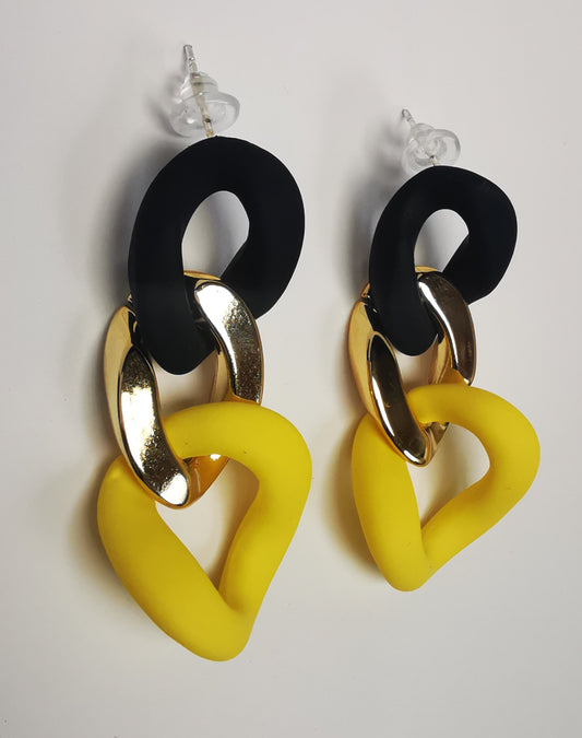 Boucles d'oreilles pendantes sur clous, posées l'une à coté de l'autre. Chacune est constituée de trois gros maillons de chaîne. Le premier en plastique noir texturé, le deuxième en métal doré, le troisième en plastique jaune texturé.