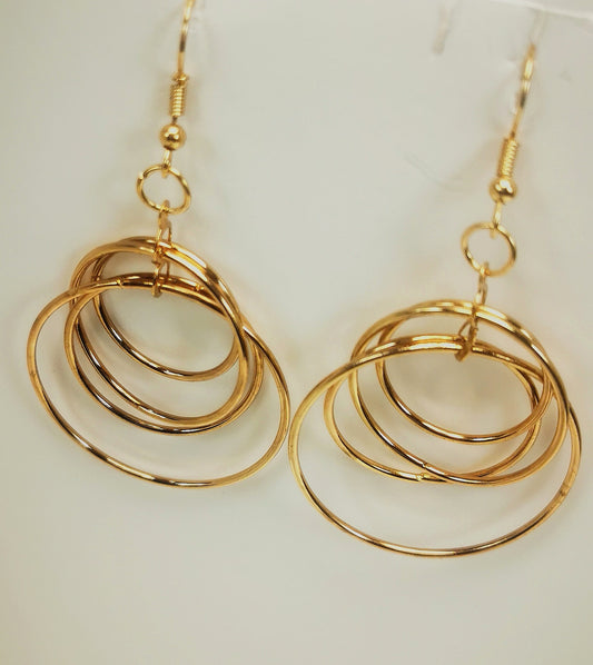Boucles d'oreilles pendantes dorées de 60mm de long. Composées de quatre anneaux imbriqués les uns dans les autres, Le tout doré est présenté en paire, accrochées à un support blanc. Vue de face.