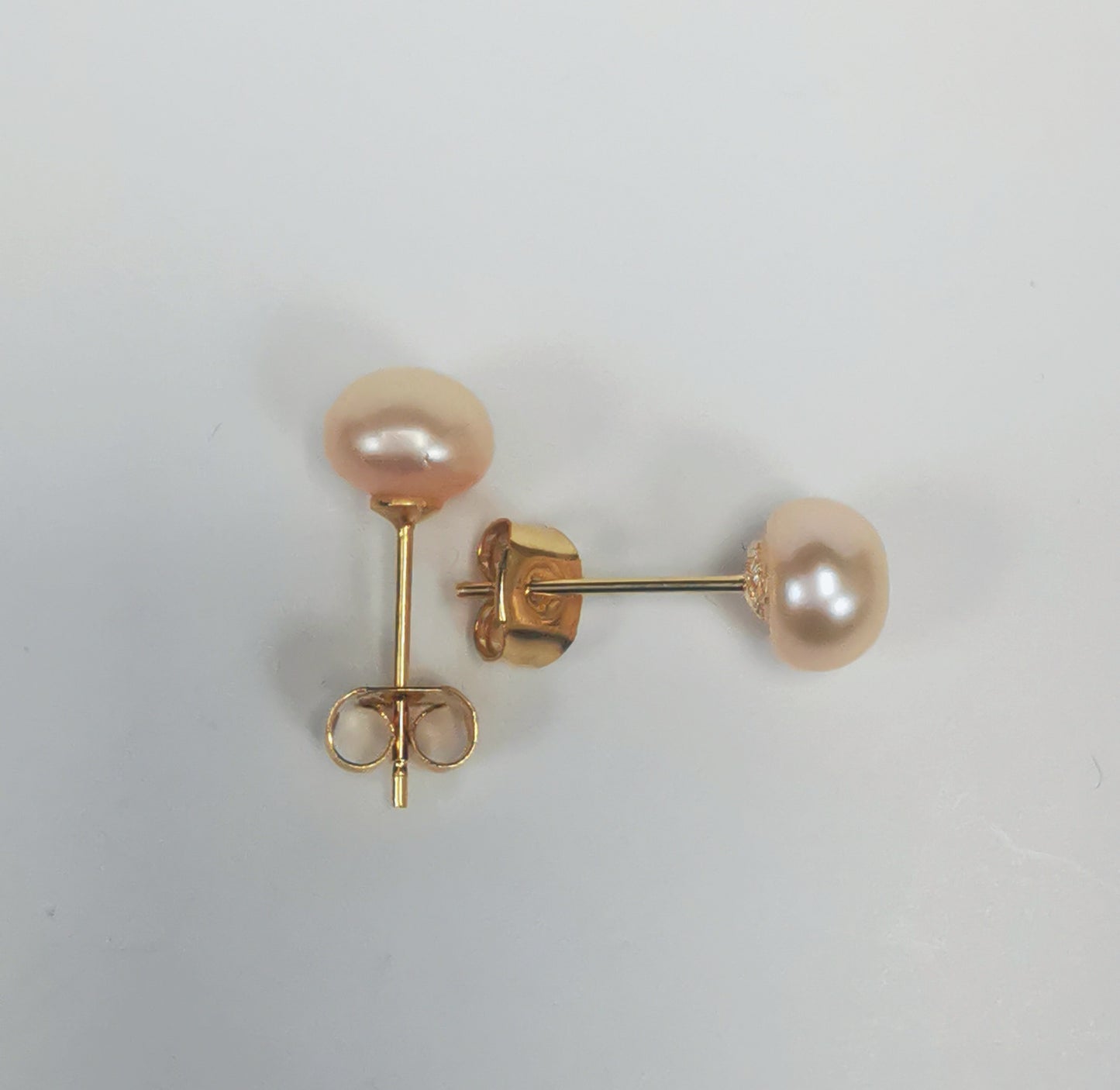 Boucles d'oreilles perles de culture rose 7mm sur clous plaqués or. Présentées perpendiculairement, boucle droite verticale, perle en haut, boucle de droite horizontale, perle à droite, sur un fond blanc. Vue de dessus.