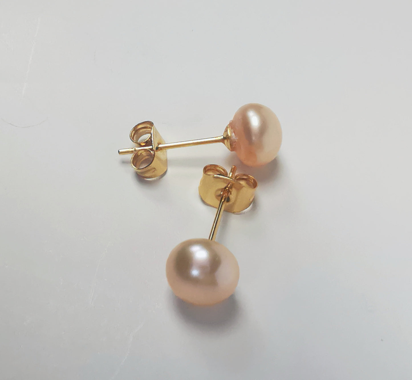 Boucles d'oreilles perles de culture rose 7mm sur clous plaqués or. Présentées perpendiculairement, boucle de dessus perle à droite, celle de dessous, verticale perle en bas sur un fond blanc. Vue de dessus.