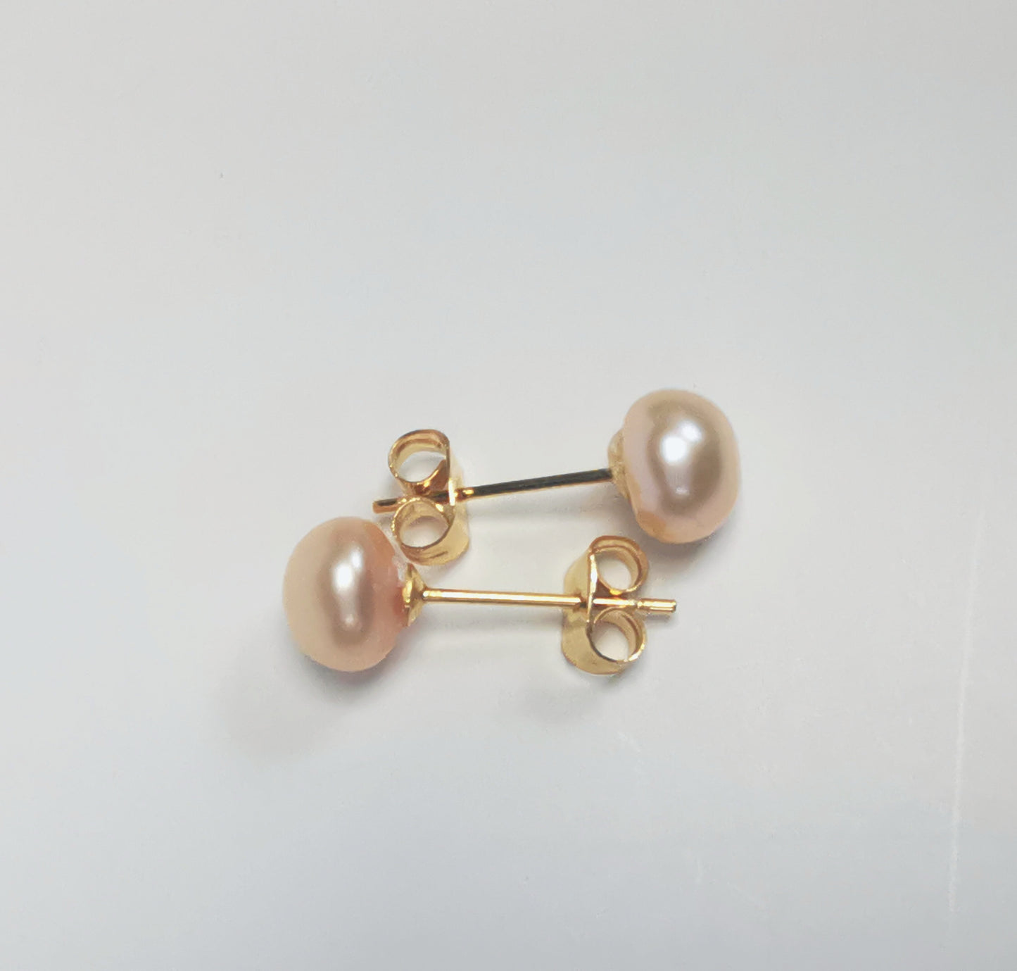 Boucles d'oreilles perles de culture rose 7mm sur clous plaqués or. Présentées cote à cote à l'horizontale, boucle de dessus perle à droite, celle de dessous, perle à gauche sur un fond blanc. Vue de dessus.