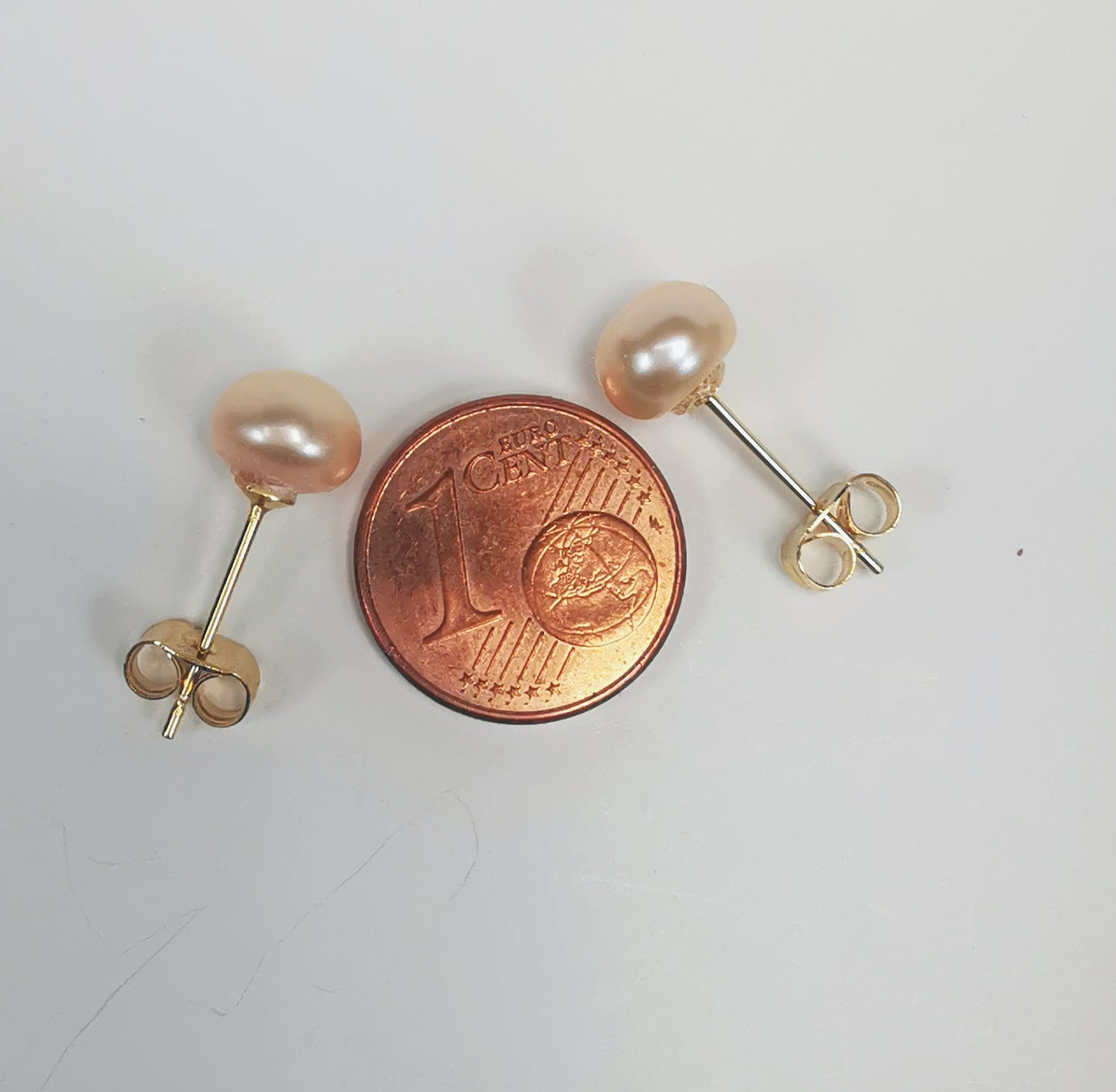 Boucles d'oreilles perles de culture rose 7mm sur clous plaqués or. Présentées perles en haut, avec une pièce de 1 centime, entre les deux, sur un fond blanc. Vue de dessus.