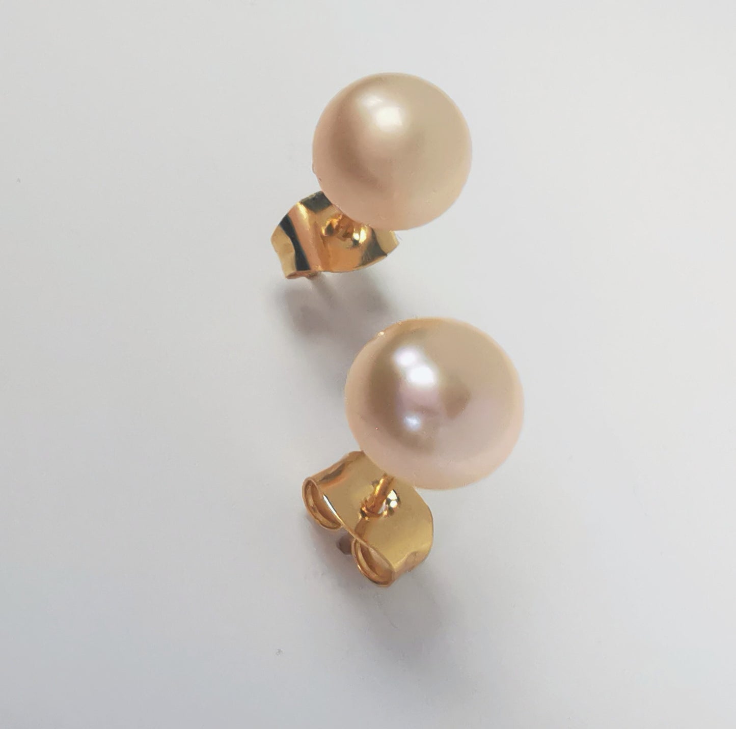 Boucles d'oreilles perles de culture rose 7mm sur clous plaqués or. Présentées l'une au dessus de l'autre plantées sur un fond blanc, inclinées vers l'arrière. Vue en très gros plan de face.