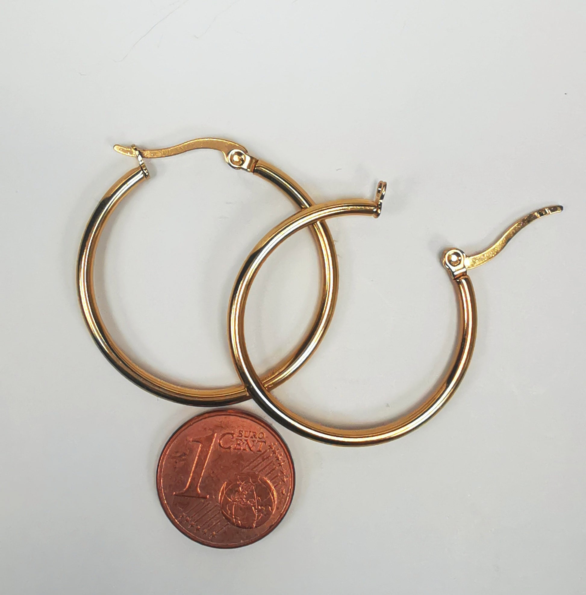 Boucles d'oreilles créoles inox plaqué or, diamètre 30mm, fil de 2mm. Présentées posées sur fond blanc, l'une sur l'autre, celle de droite fermoir ouvert, au dessus d'une pièce de 1 centime. Vue de dessus.