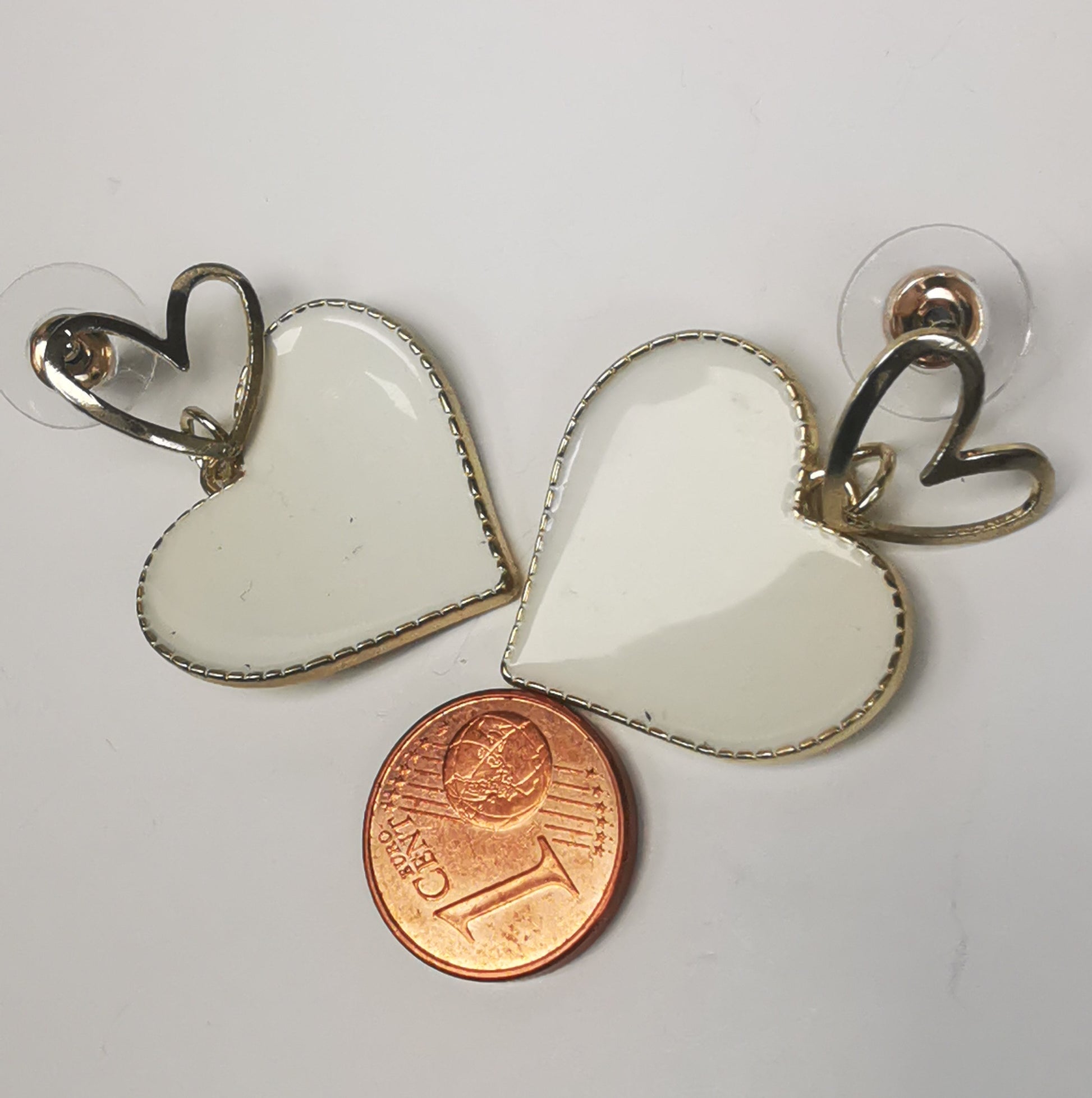 Paire de boucles d'oreilles, un petit coeur sur clou sur lequel pend un coeur blanc sur entourage et fond doré. Les deux à coté d'une pièce de 1 centime.