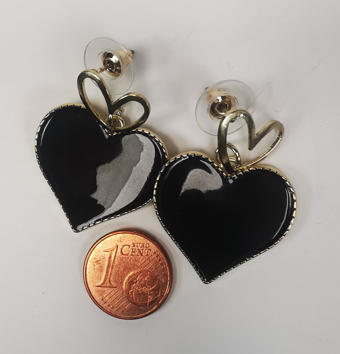 Paire de boucles d'oreilles, un petit coeur doré sur clou sur lequel pend un coeur noir sur entourage et fond doré posée à coté d'une pièce de 1 centime.