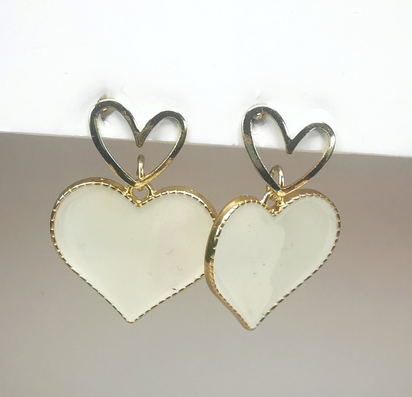 Paire de boucles d'oreilles, un petit coeur sur clou sur lequel pend un coeur blanc sur entourage et fond doré; Les deux  sont pendantes , accrochées à un clou doré.