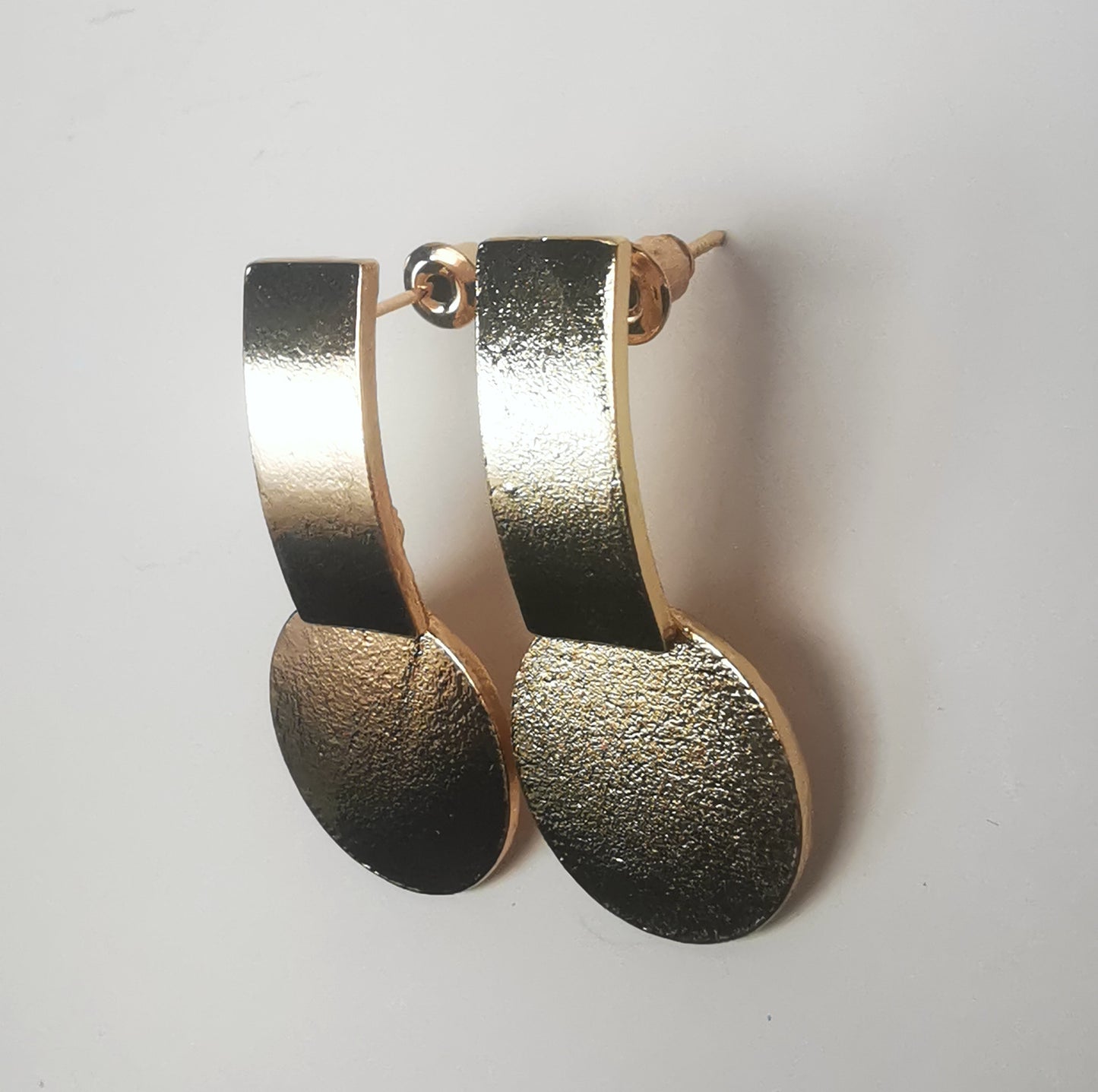 Boucles d'oreilles sur clous en métal doré. Un premier rectangle vertical est fixé au clou. En dessus est suspendu un disque. Le tout est texturé. Présentées cote à cote, posées sur un fond blanc. Vue depuis la droite.