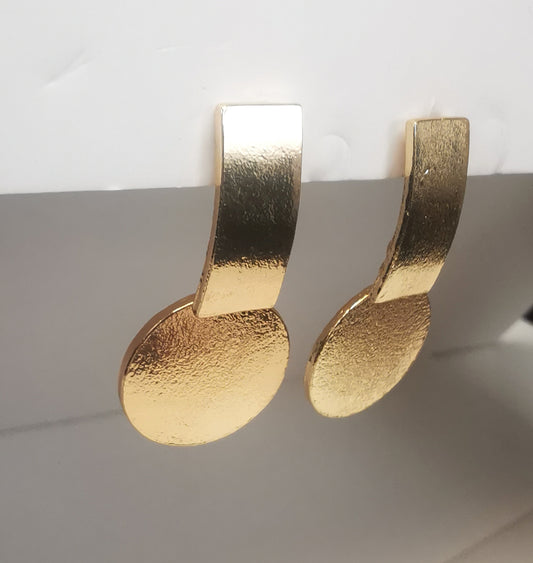 Boucles d'oreilles sur clous en métal doré. Un premier rectangle vertical est fixé au clou. En dessus est suspendu un disque. Le tout est texturé. Présentées cote à cote, accrochées sous un support blanc. Vue depuis la gauche.